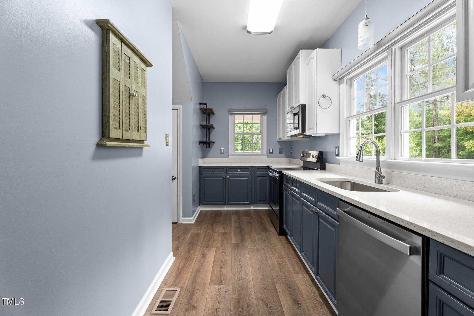Modern kitchen with blue lower cabinets, white upper cabinets, stainless steel appliances, light wood floors, large windows, and light blue walls. Natural light fills the space, creating an airy feel.