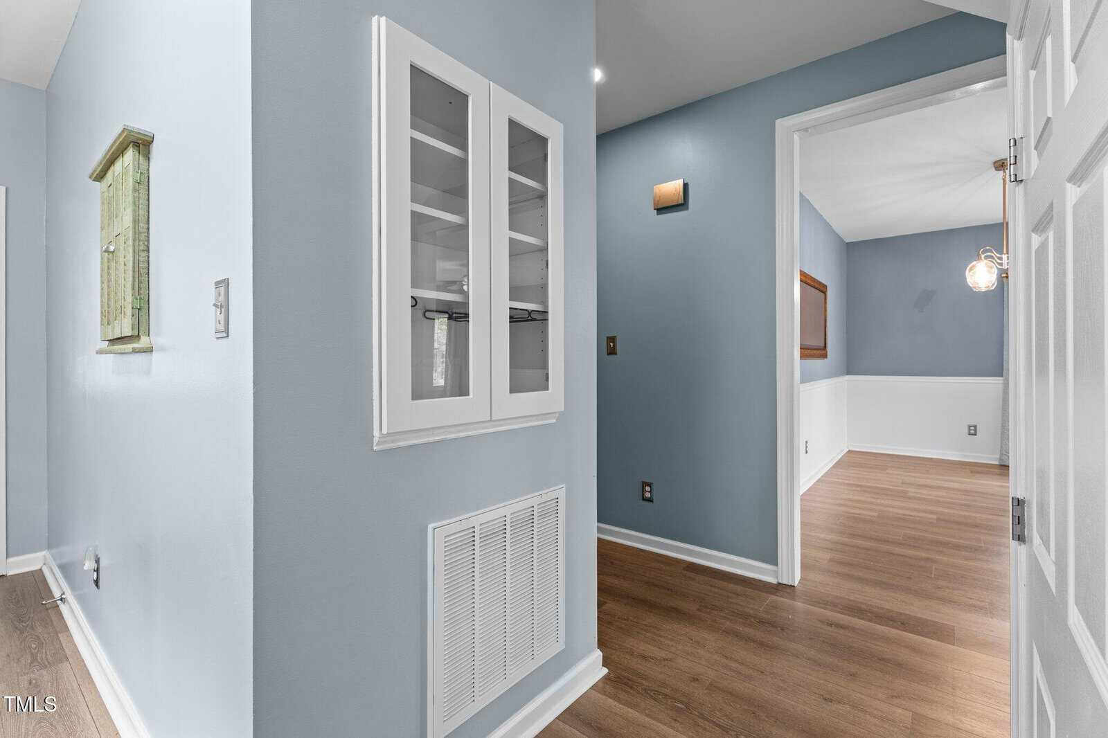 A hallway with light blue walls, wood flooring, a built-in white cabinet with glass doors, and a vent cover below. An adjacent room has white wainscoting and a modern light fixture.