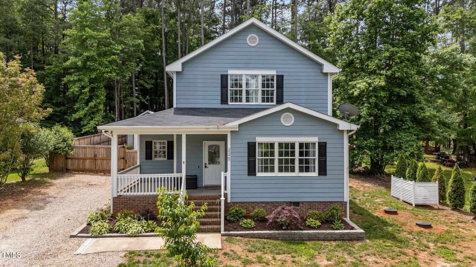 A charming two-story blue house with white trim on 225 Sacred Fire Road, Louisburg, featuring a covered front porch, surrounded by trees and greenery. A gravel driveway leads to the side and a small fenced area sits on the right.