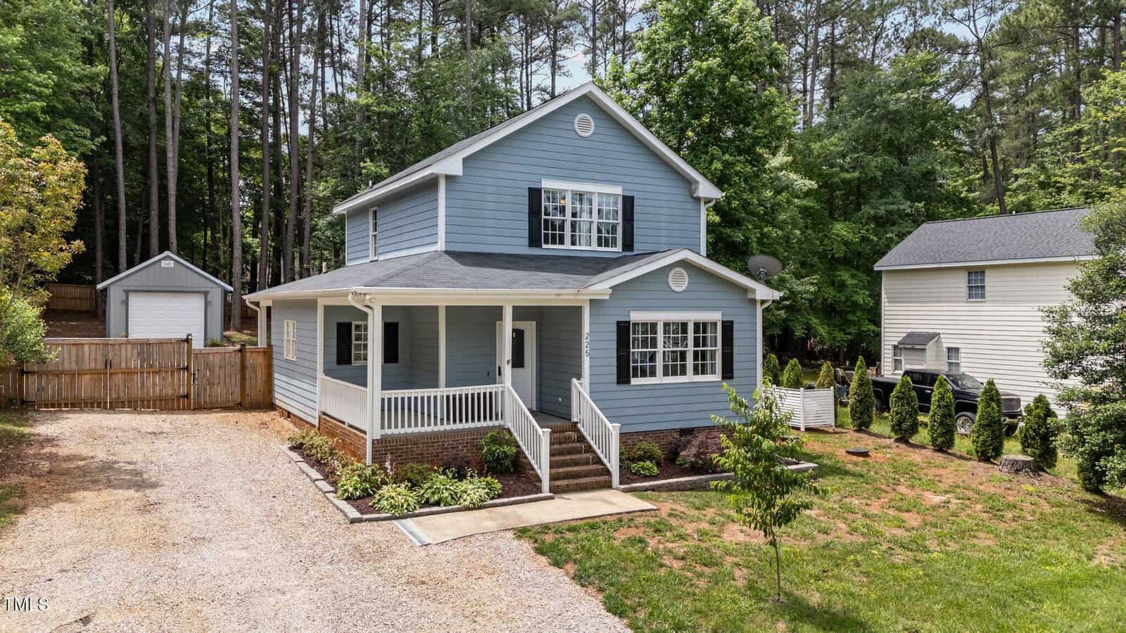 A two-story blue house with white trim, a covered front porch, and a gravel driveway. The yard is landscaped with grass and shrubs, and there are tall trees in the background. A detached garage is visible in back.