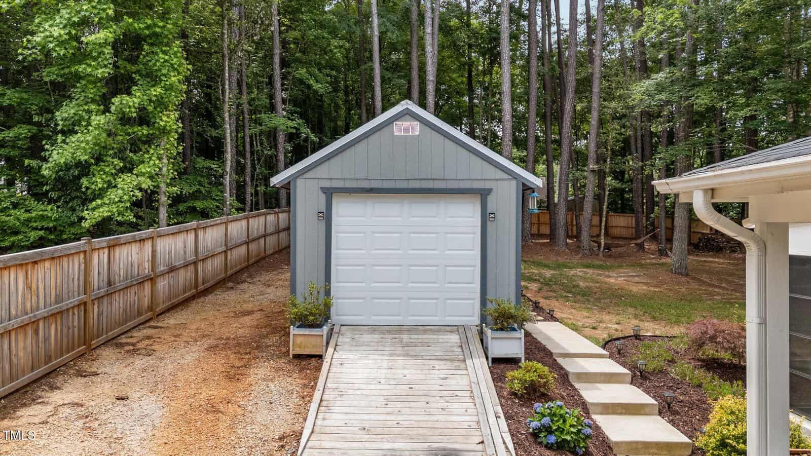 A small blue-gray shed with a white garage door sits at the end of a wooden ramp, surrounded by trees, a wooden fence, and landscaped gardens beside a gravel path.