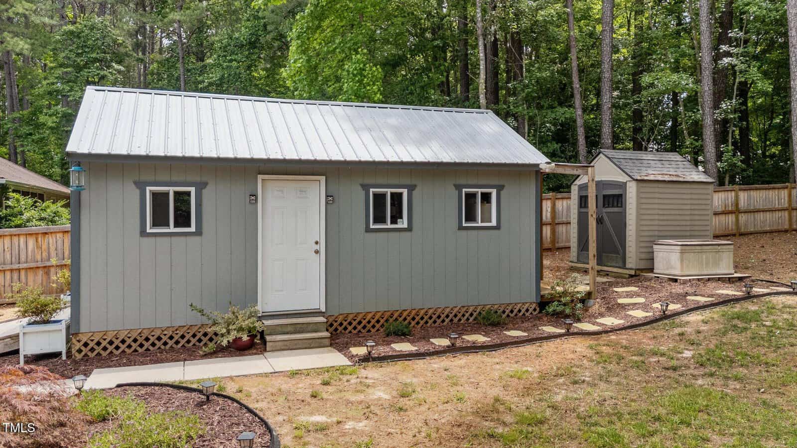 Gray shed with a metal roof in a fenced backyard surrounded by trees. A paved path leads to the door, with small plants along the edge. A smaller shed and a stepping-stone path are visible to the right.