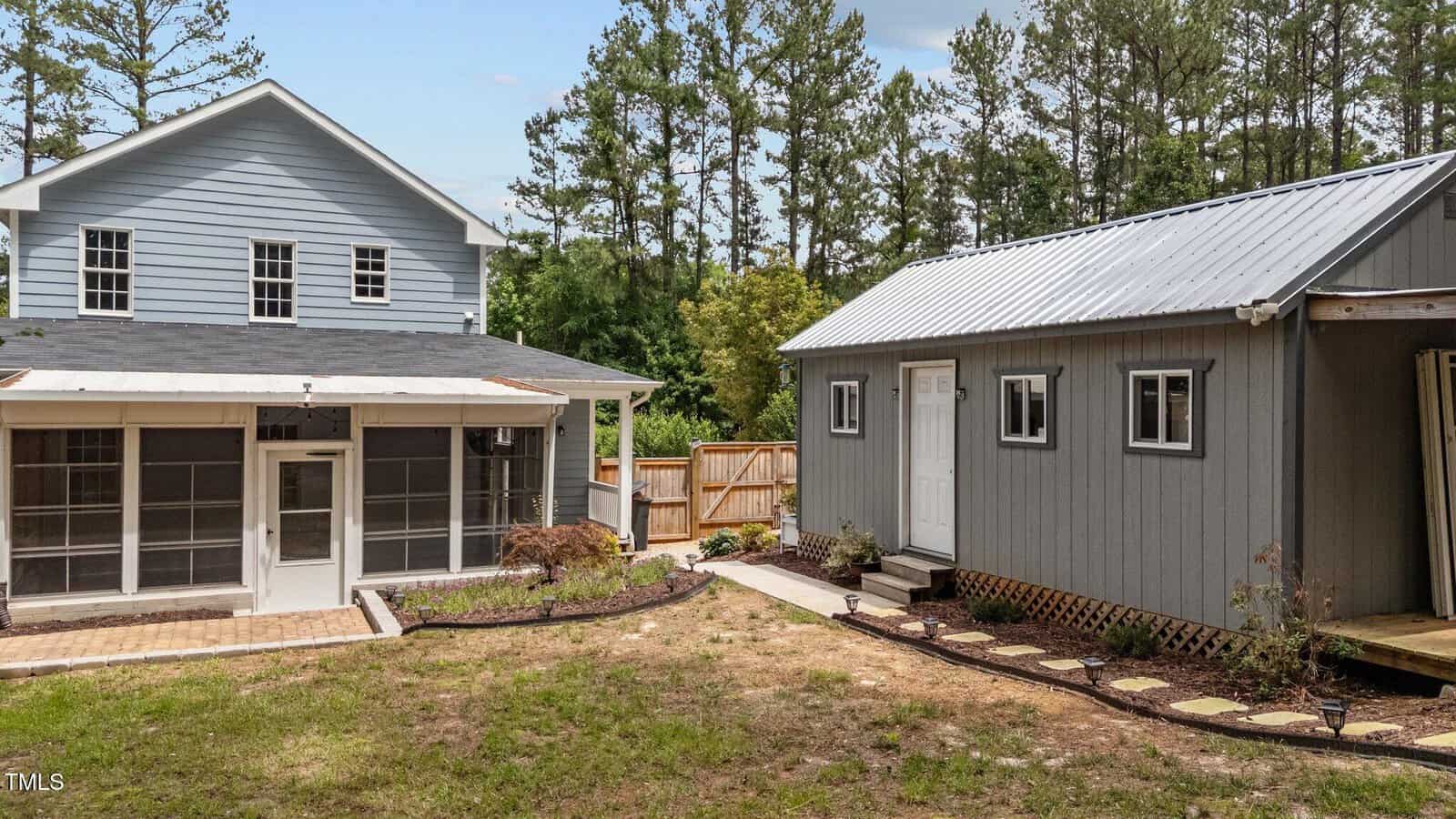 A gray two-story house with a screened-in porch sits beside a smaller gray outbuilding with a metal roof, surrounded by a fenced yard and trees in the background.