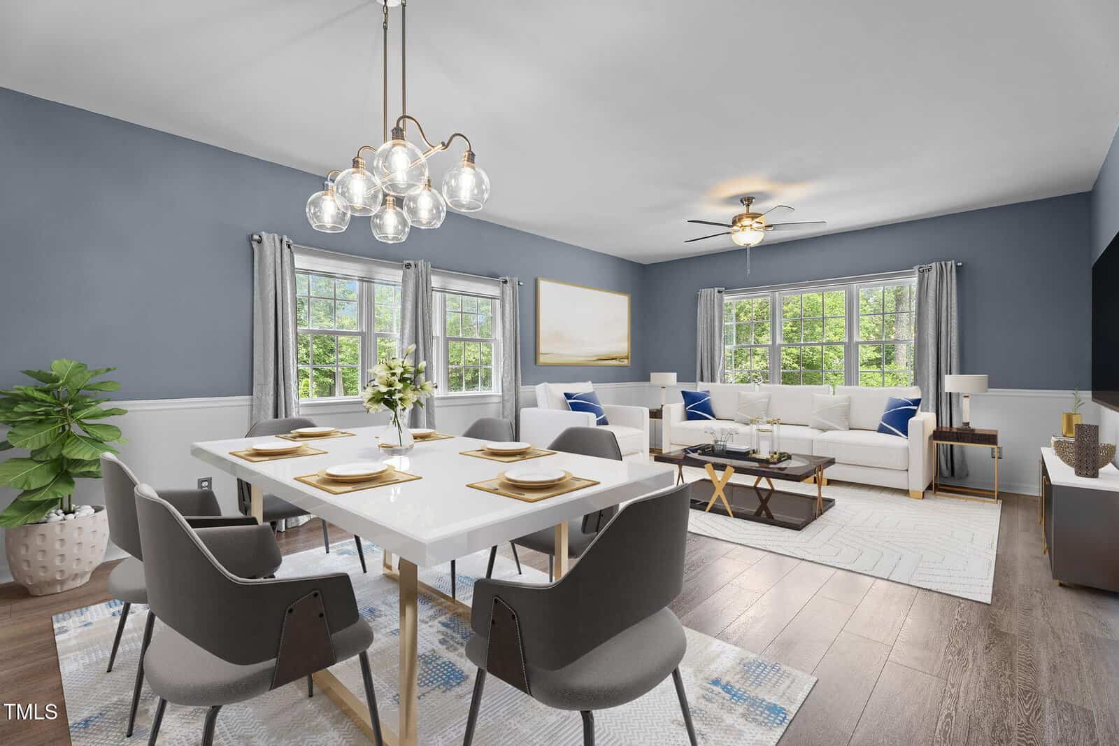 Modern dining and living room with blue-gray walls, white trim, wood floors, large windows, stylish chandelier, dining table set for four, white sofa, and decorative accents; space is bright and airy with natural light.
