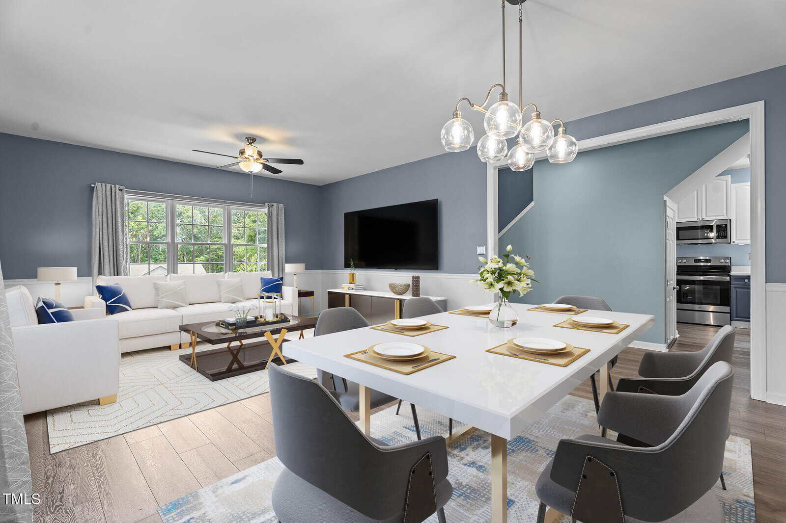 A modern open-concept living and dining area with blue-gray walls, a white sectional sofa, a large TV, a white dining table set for six, contemporary chairs, and pendant lighting. Large windows provide natural light.
