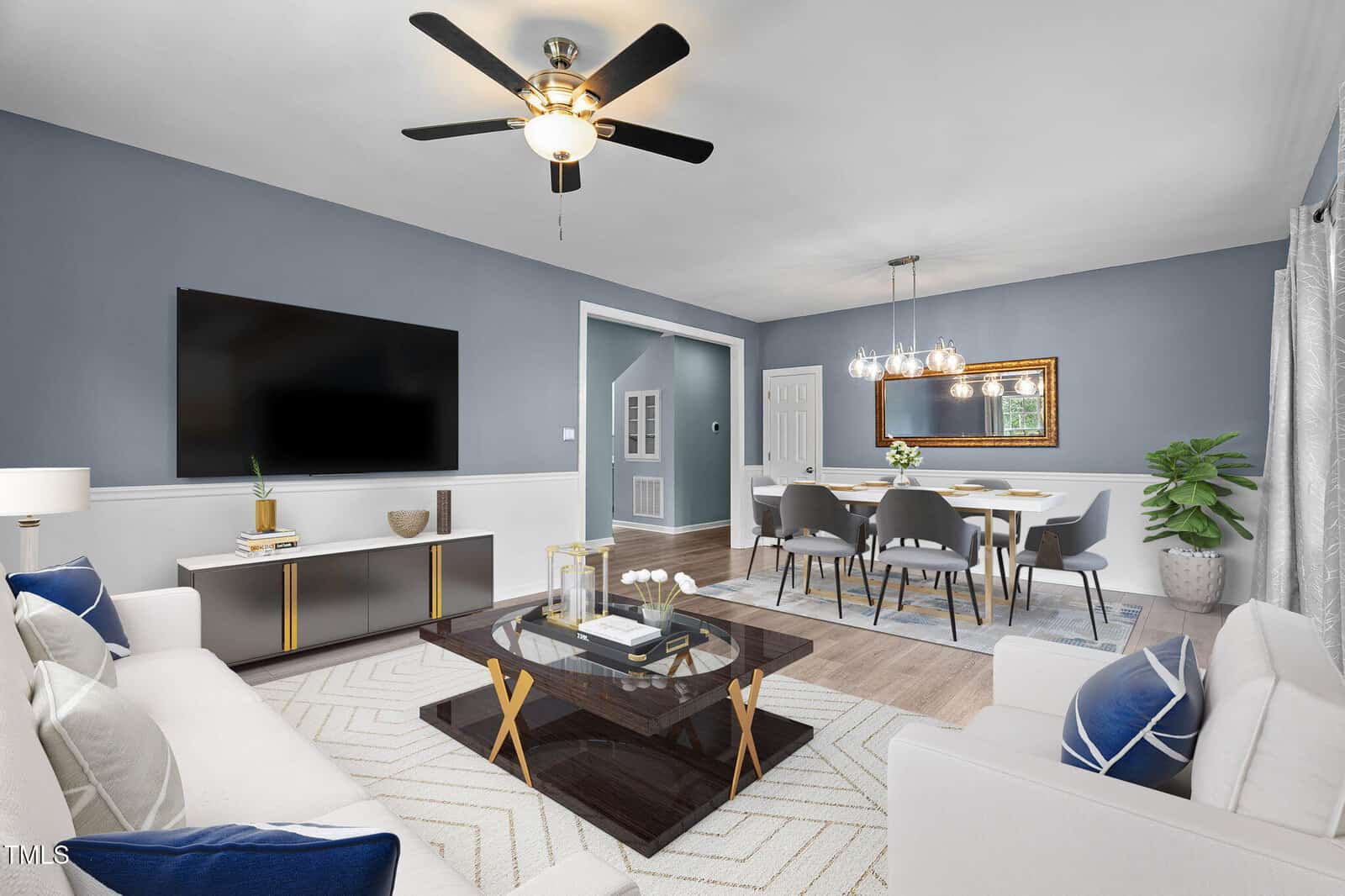 A modern living and dining room with blue walls, white furniture, a large wall-mounted TV, a glass coffee table, a ceiling fan, and a dining table set for six under a chandelier. Light wood floors and a potted plant add a fresh touch.