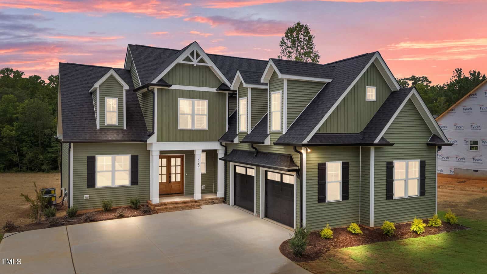 A two-story green house with white trim and black shutters, featuring double front doors and a two-car garage, is illuminated at sunset with a wooded background and a neighboring house under construction.