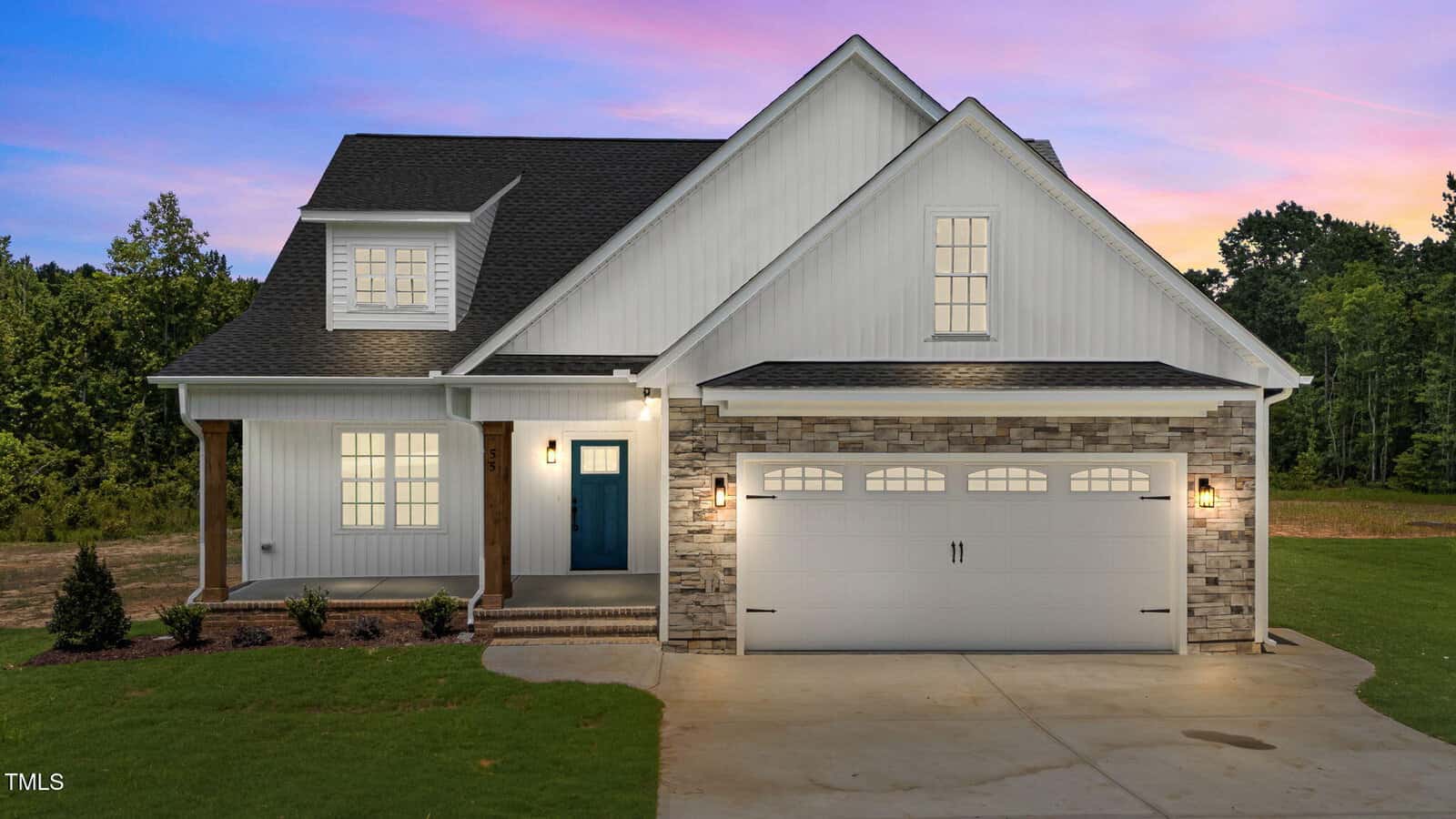 Two-story modern farmhouse with white siding, brick accents, a blue front door, and a two-car garage. Exterior lights are on, and trees and a sunset sky are in the background.