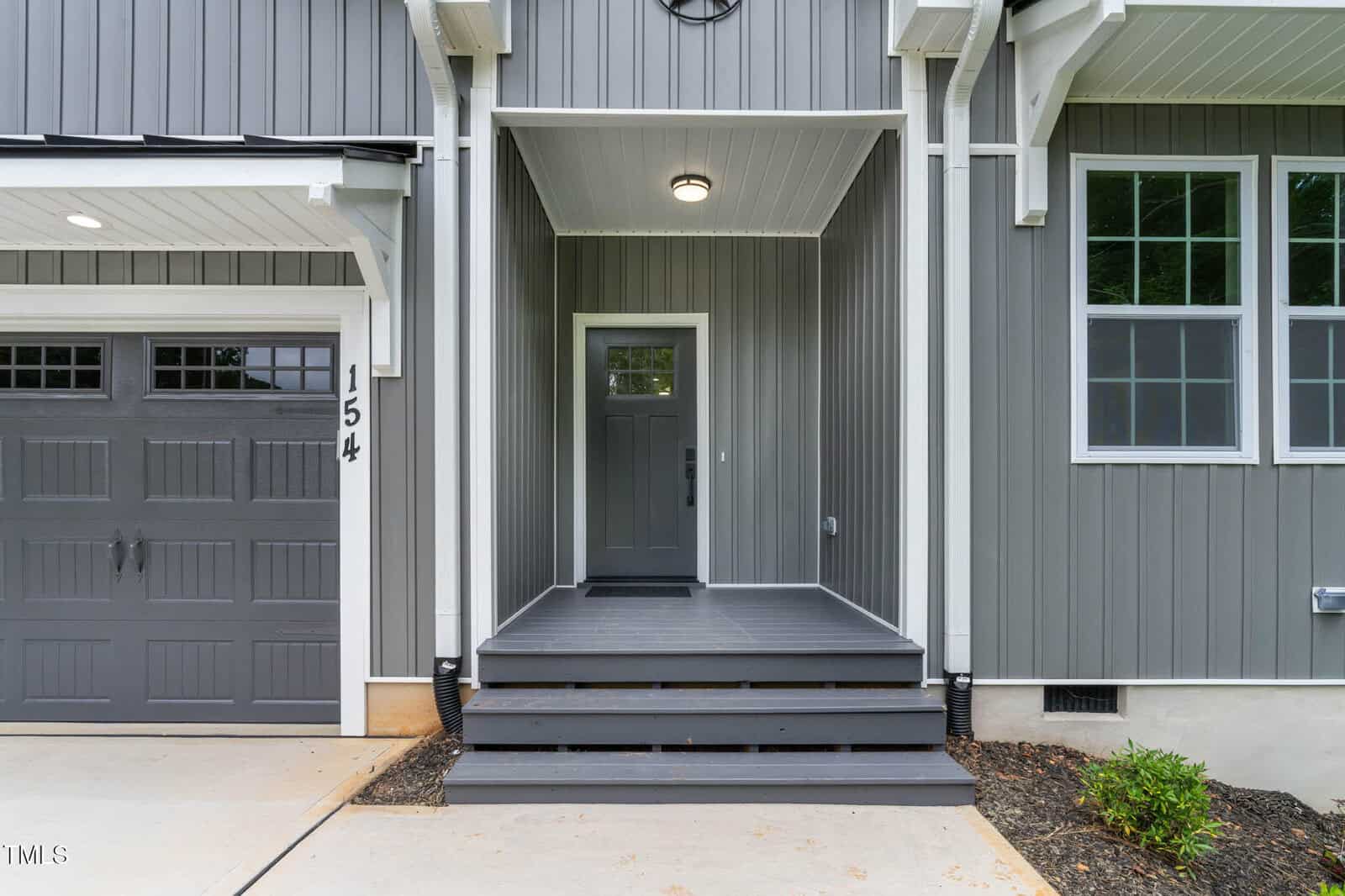 A gray front door with a covered porch, steps, and white trim, next to a gray garage door with the number 154, on a modern house with vertical siding.