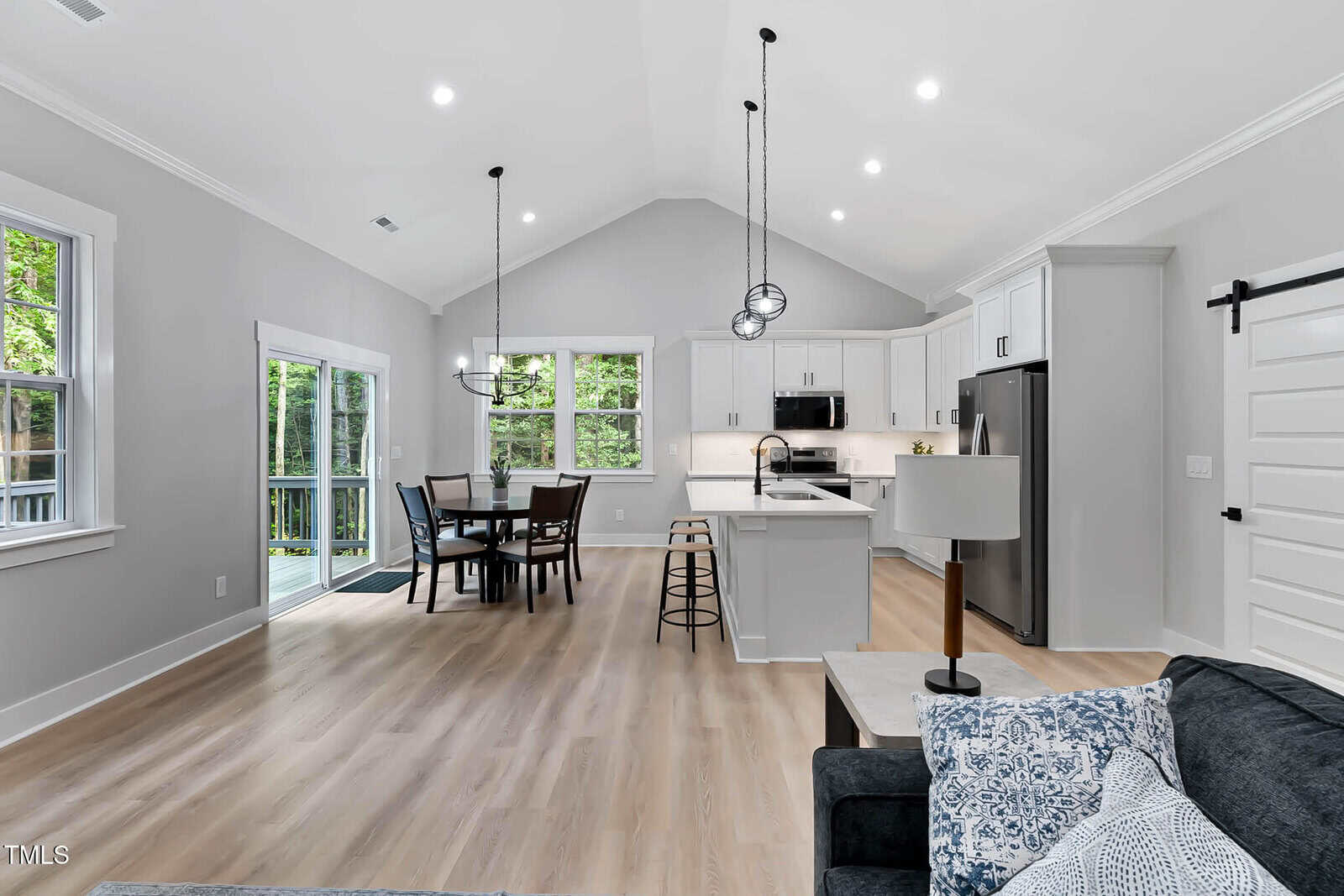 Modern open-concept kitchen and dining area with light wood floors, white cabinetry, stainless steel appliances, and a center island with barstools. Large windows and glass doors overlook a green outdoor space.