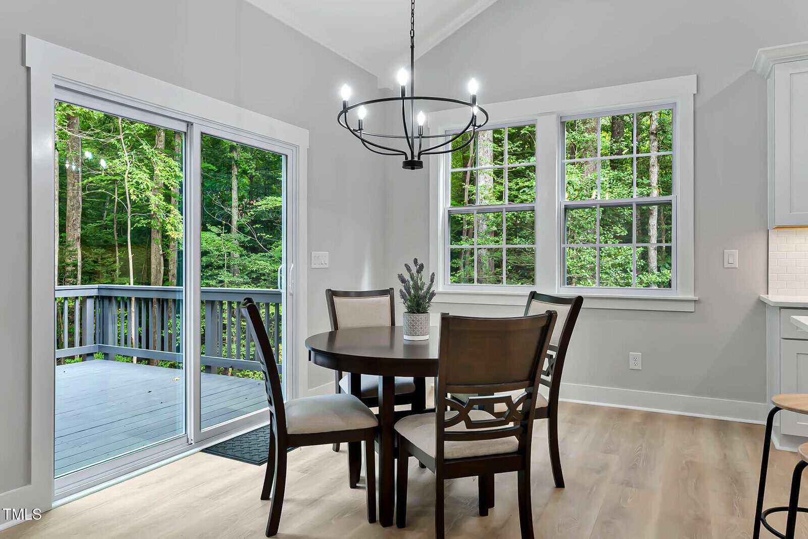 A round dining table with four chairs sits near sliding glass doors and a window, overlooking a wooded deck. The room features light wood flooring and a modern black chandelier.