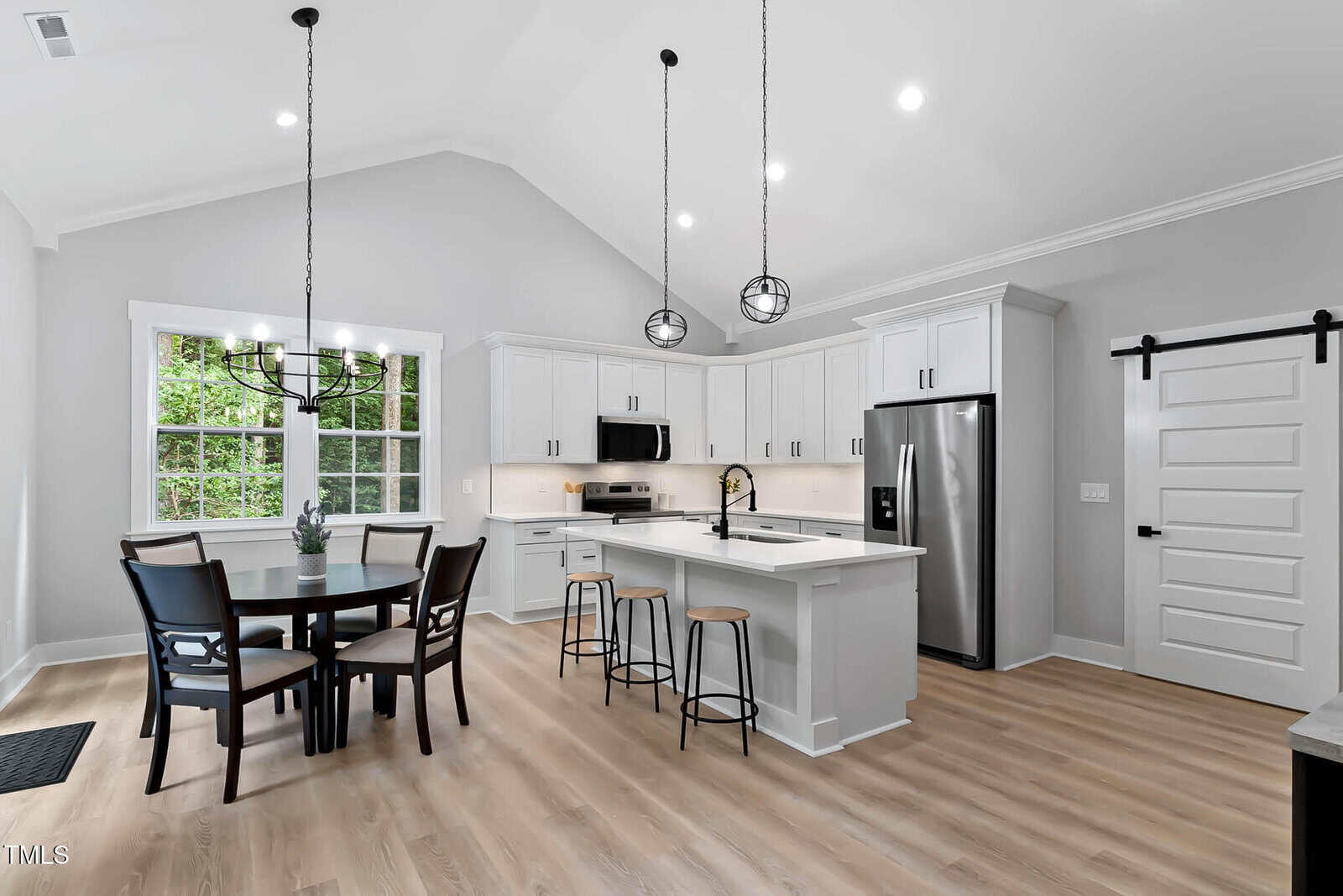 Modern kitchen with white cabinets, stainless steel appliances, an island with three stools, a round dining table with four chairs by a window, and two pendant lights hanging from a vaulted ceiling.