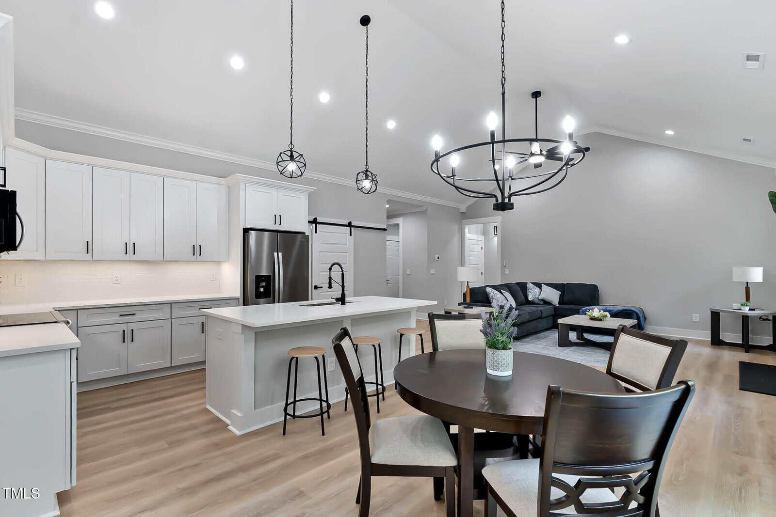 Modern open-concept kitchen and living area with white cabinets, stainless steel appliances, island with stools, round dining table, and sectional sofa. Light wood flooring and recessed lighting are featured throughout.