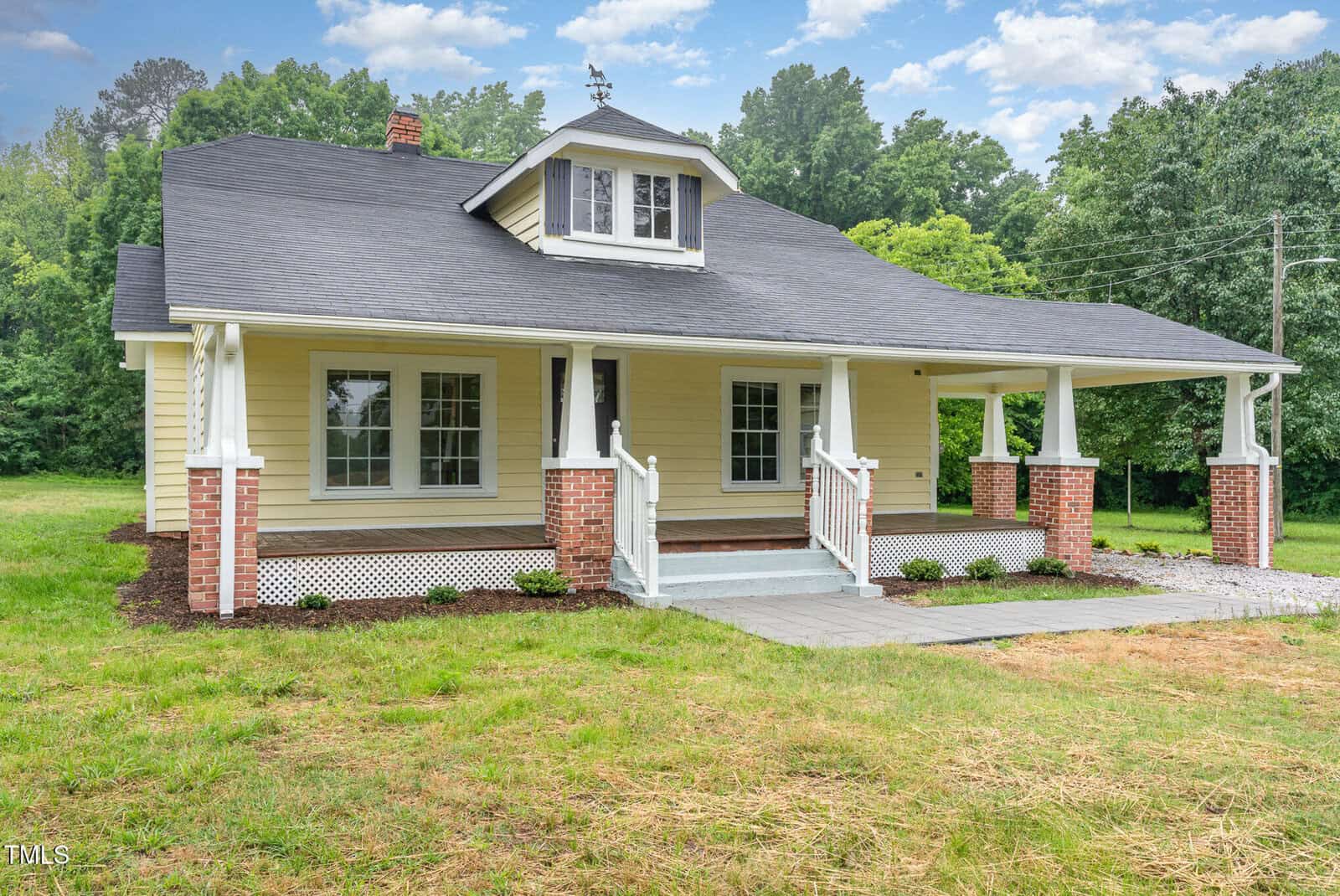 A charming, yellow single-story house with a covered front porch, white trim, brick columns, and a dark roof, surrounded by green grass and trees under a partly cloudy sky.