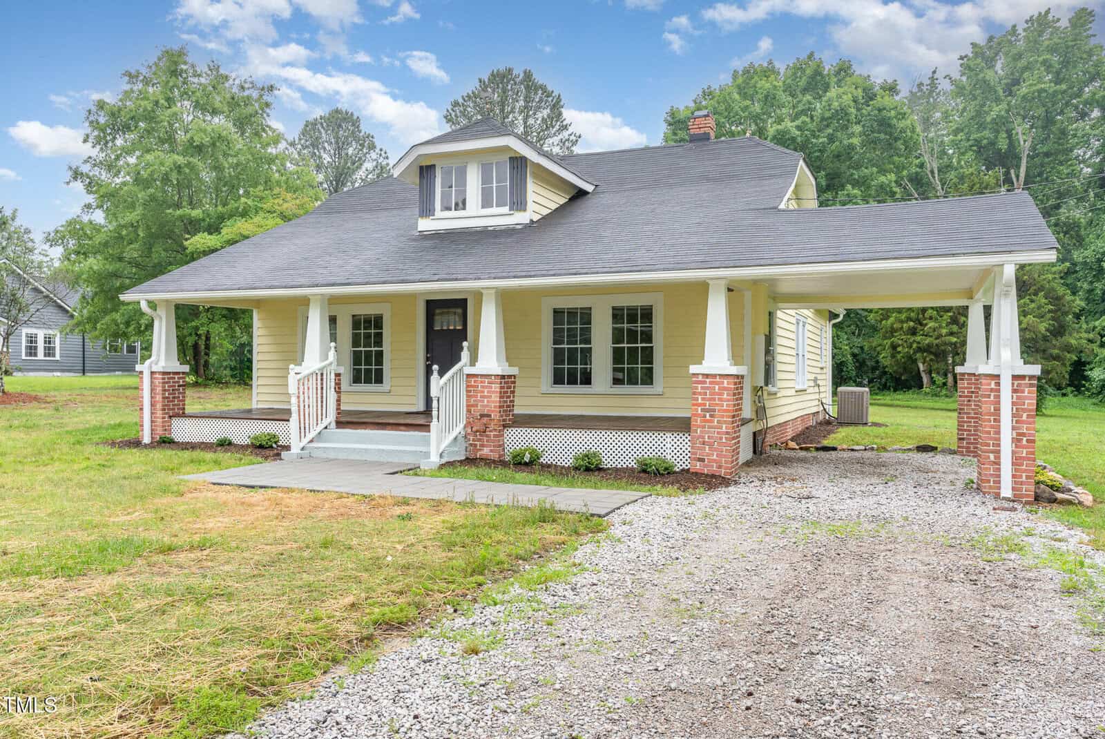 A yellow house with white trim, a covered front porch, and a gabled dormer. The house has brick pillars, a carport on the right, and is surrounded by grass, trees, and a gravel driveway under a partly cloudy sky.