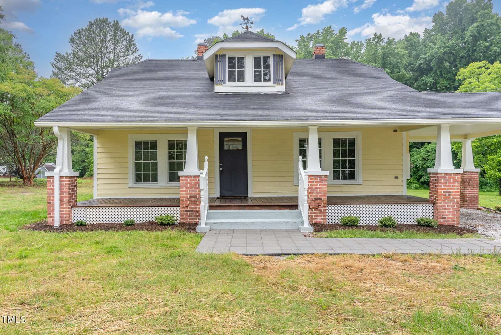 A yellow Craftsman-style house with a covered front porch, white railings and trim, red brick pillars, a black front door, and a dormer window, surrounded by grass and trees under a partly cloudy sky.
