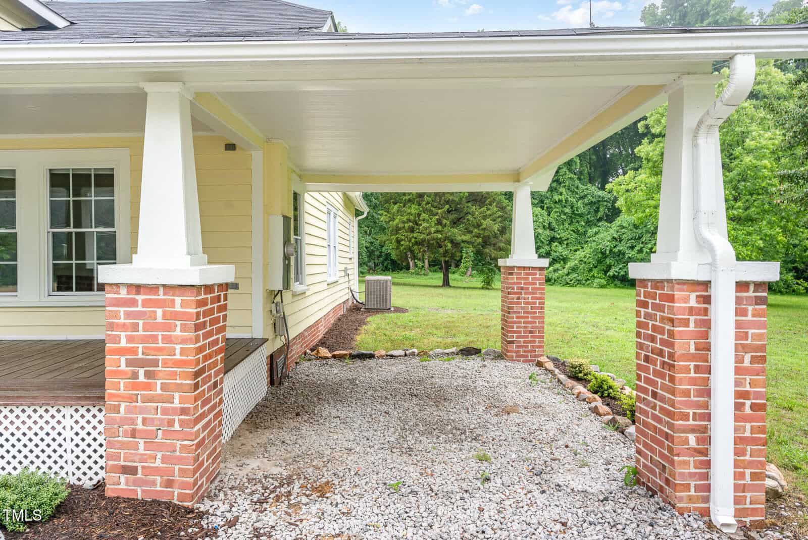 A covered carport with brick and white columns beside a yellow house, gravel flooring, and green lawn with trees in the background.