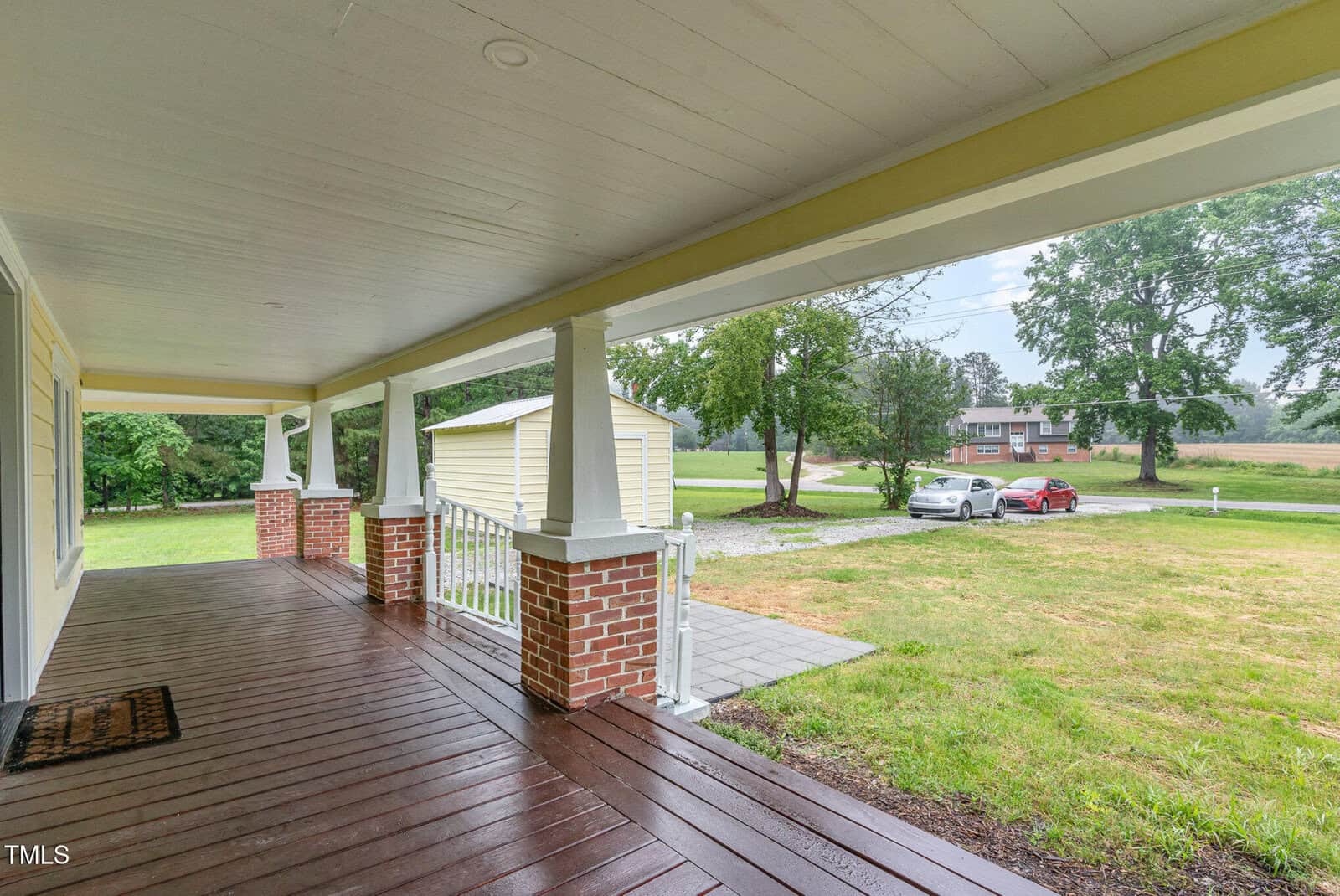 Covered wooden front porch with white railing and brick columns overlooks a grassy yard with a driveway, detached garage, and several cars parked near trees and a house in the distance.