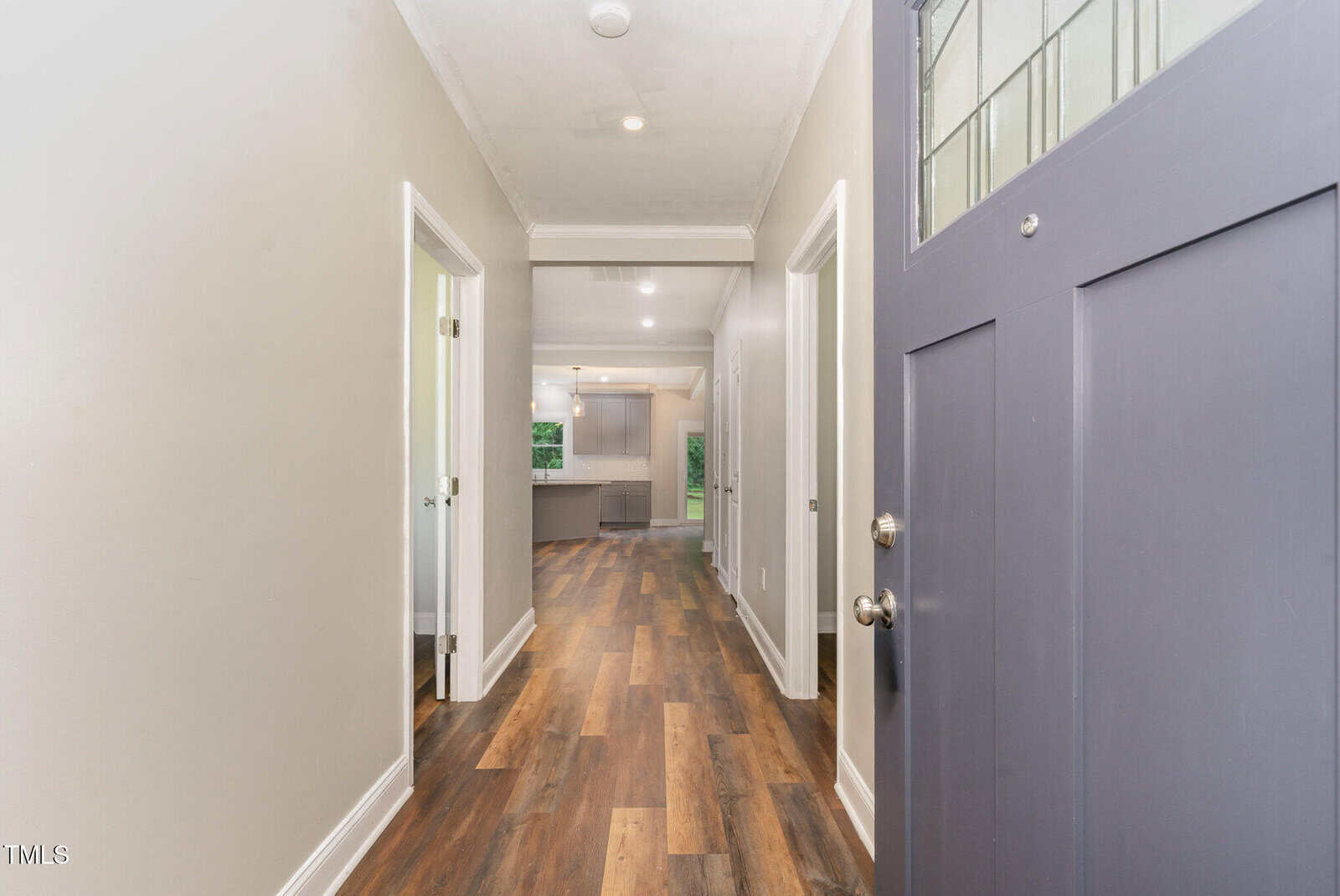 View from an open front door looking down a hallway with wood flooring, neutral-colored walls, and doorways on the left, leading to a bright kitchen area at the end of the hall.