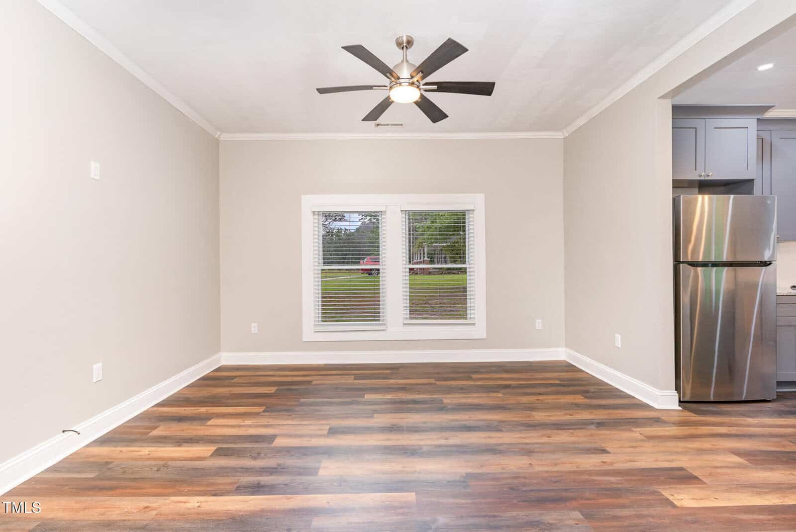 Unfurnished room with light beige walls, wood-look flooring, white trim, a ceiling fan with lights, a window with blinds, and a partial view of a kitchen with a stainless steel refrigerator.
