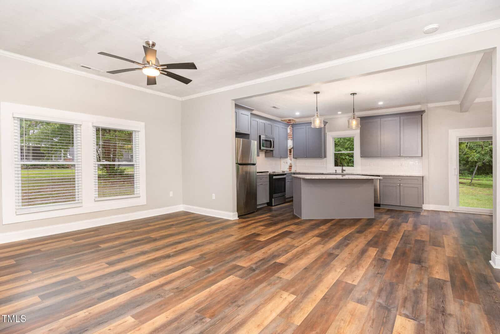 Open-concept room with wood-look flooring, a ceiling fan, and a modern kitchen featuring gray cabinets, stainless steel appliances, pendant lights, and large windows overlooking a green yard.
