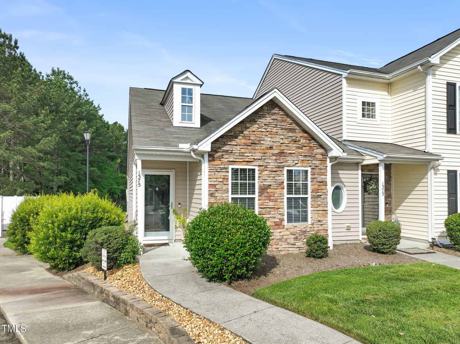 A two-story townhouse at 1375 Quail Circle in Creedmoor features beige siding, a partial stone facade, white front door, and dormer window. The home is surrounded by greenery, shrubs, a sidewalk, and small lawn under a clear sky.