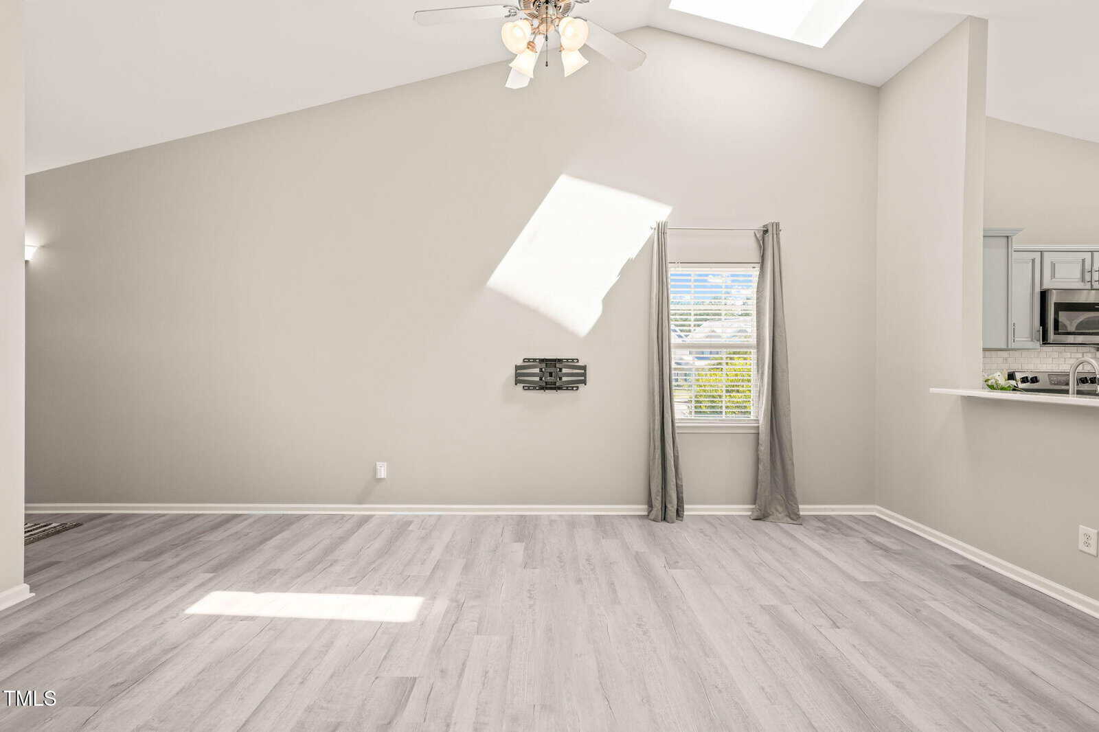 A bright, empty living room with light wood flooring, light gray walls, a ceiling fan, wall-mounted TV bracket, small window with gray curtains, and a partial view of a kitchen with white countertops.