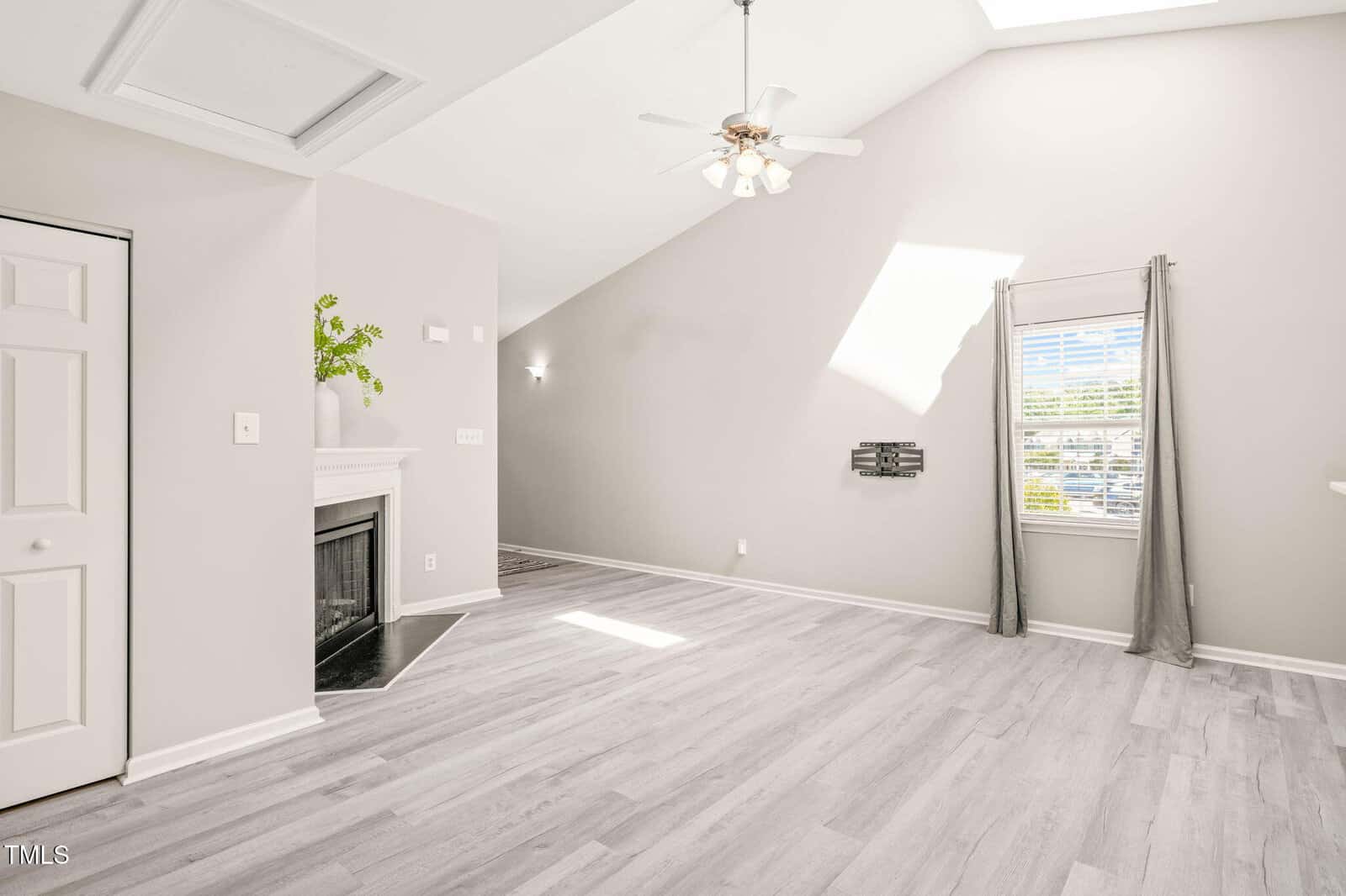 A spacious, empty living room with light-colored wood flooring, a corner fireplace with a plant on the mantle, a ceiling fan, a window with gray curtains, and sunlight streaming through a skylight.