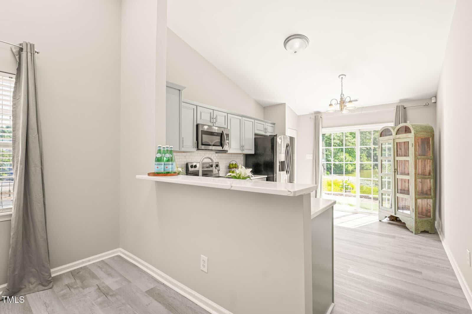 A modern kitchen with light gray cabinets, stainless steel appliances, and a white countertop, opening to a bright living area with large windows and a wooden cabinet near sliding glass doors.