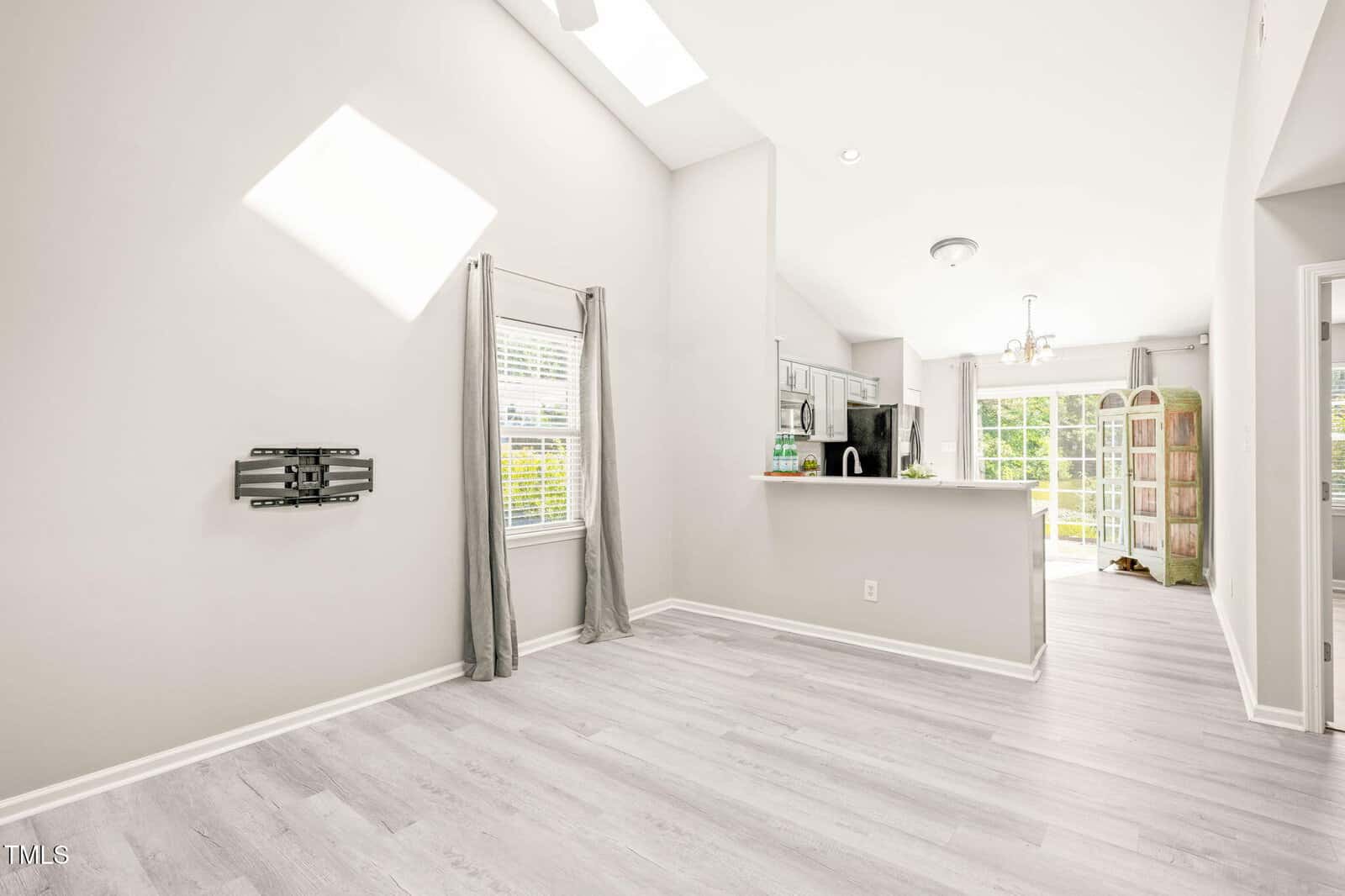 A bright, empty room with light gray wood flooring, light gray walls, a window with gray curtains, a TV wall mount, and an open view into a kitchen with stainless appliances and a sunlit dining area.