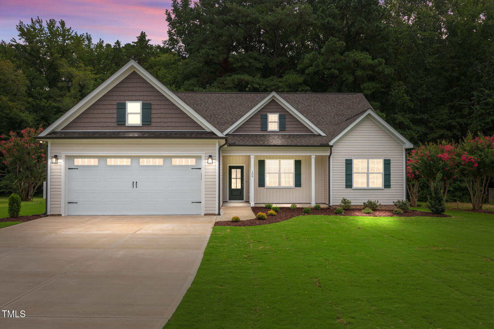 Single-story suburban house with gray siding, a double garage, front porch, and well-maintained lawn, photographed at dusk with exterior lights on and trees in the background.