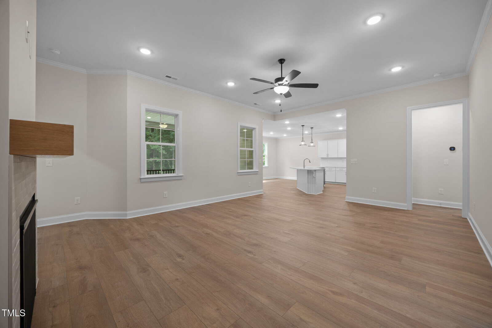 Spacious living area with light wood flooring, recessed lighting, ceiling fan, and open layout leading to a modern kitchen with white cabinetry and island. Large windows provide natural light.
