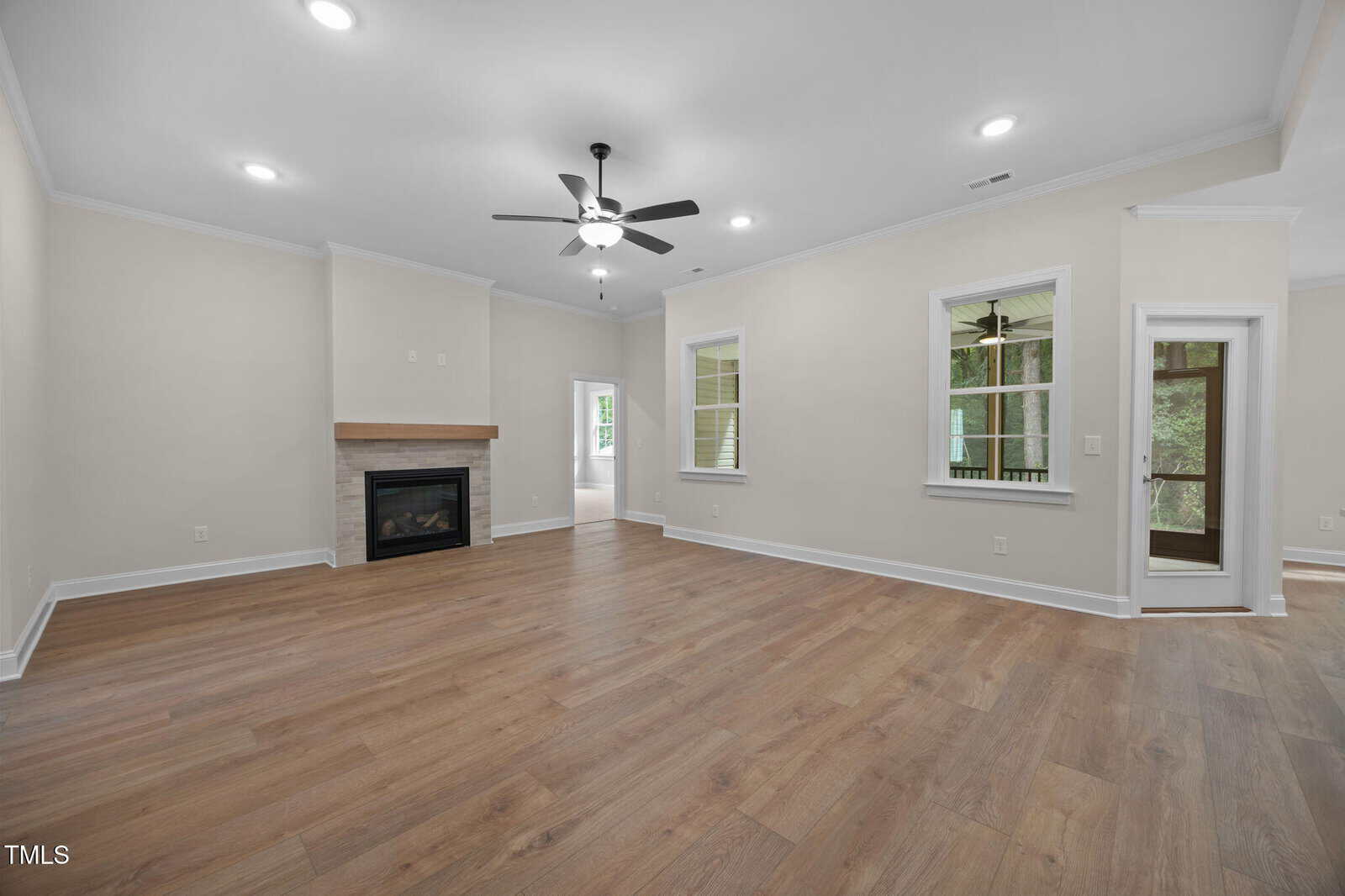 Spacious, empty living room with light-colored walls, wood flooring, a ceiling fan with lights, a corner fireplace, several windows, and a glass door leading to an enclosed porch or sunroom.