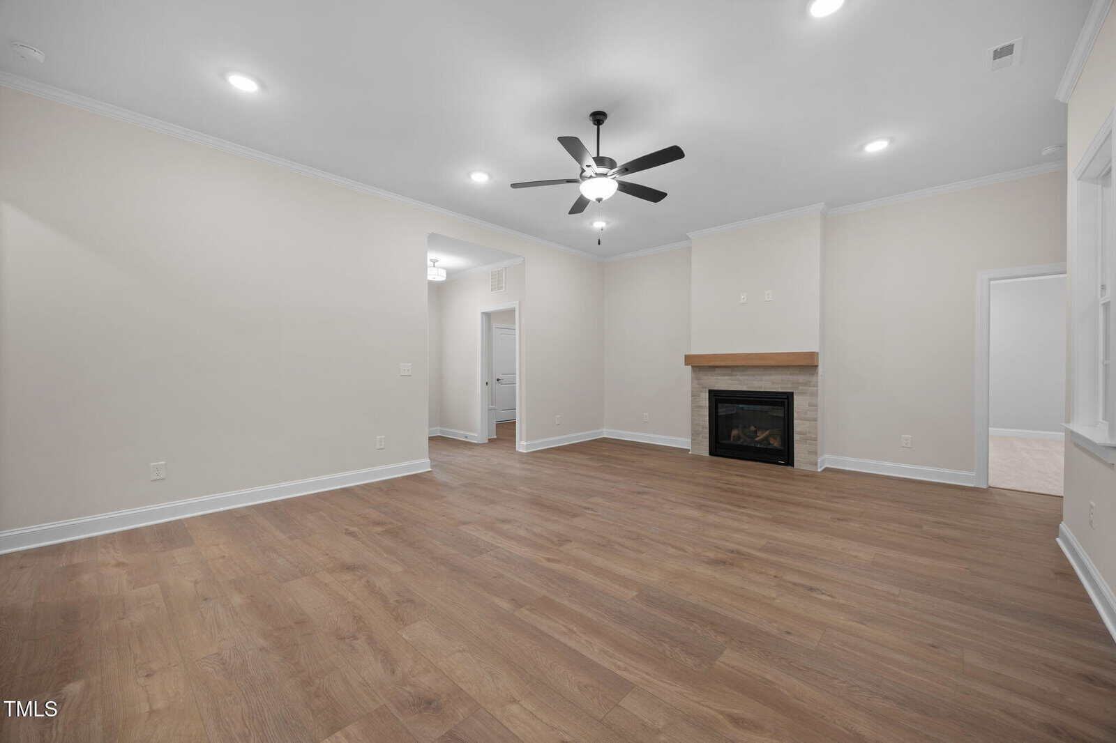 A spacious living room with light beige walls, wood flooring, a ceiling fan with lights, recessed ceiling lights, and a corner fireplace with a white mantel. Doorways lead to adjacent rooms.