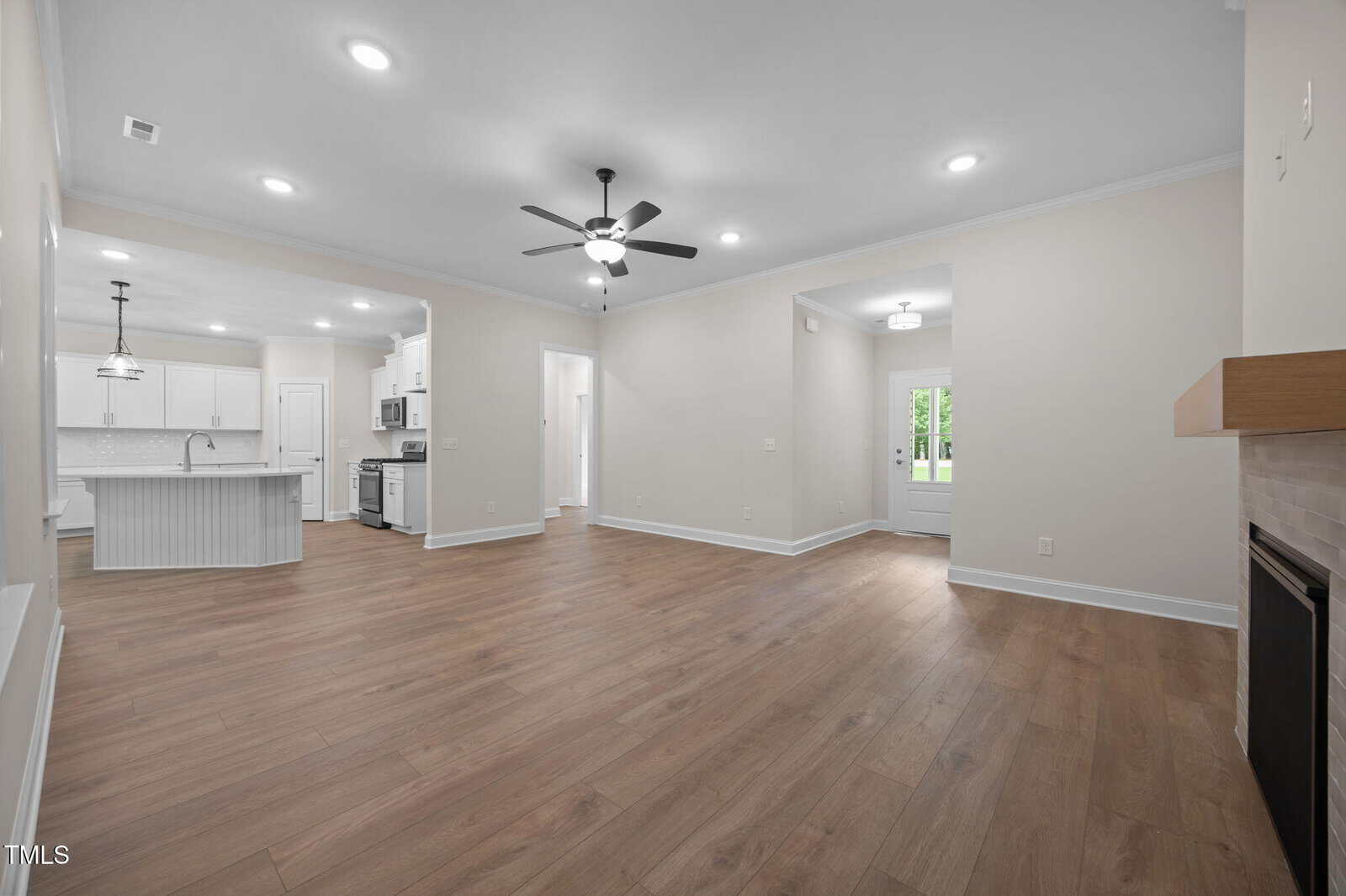 Spacious open-plan living area with wood flooring, ceiling fans, recessed lighting, a fireplace, and a view into a modern white kitchen with an island. French door leads to the outside.