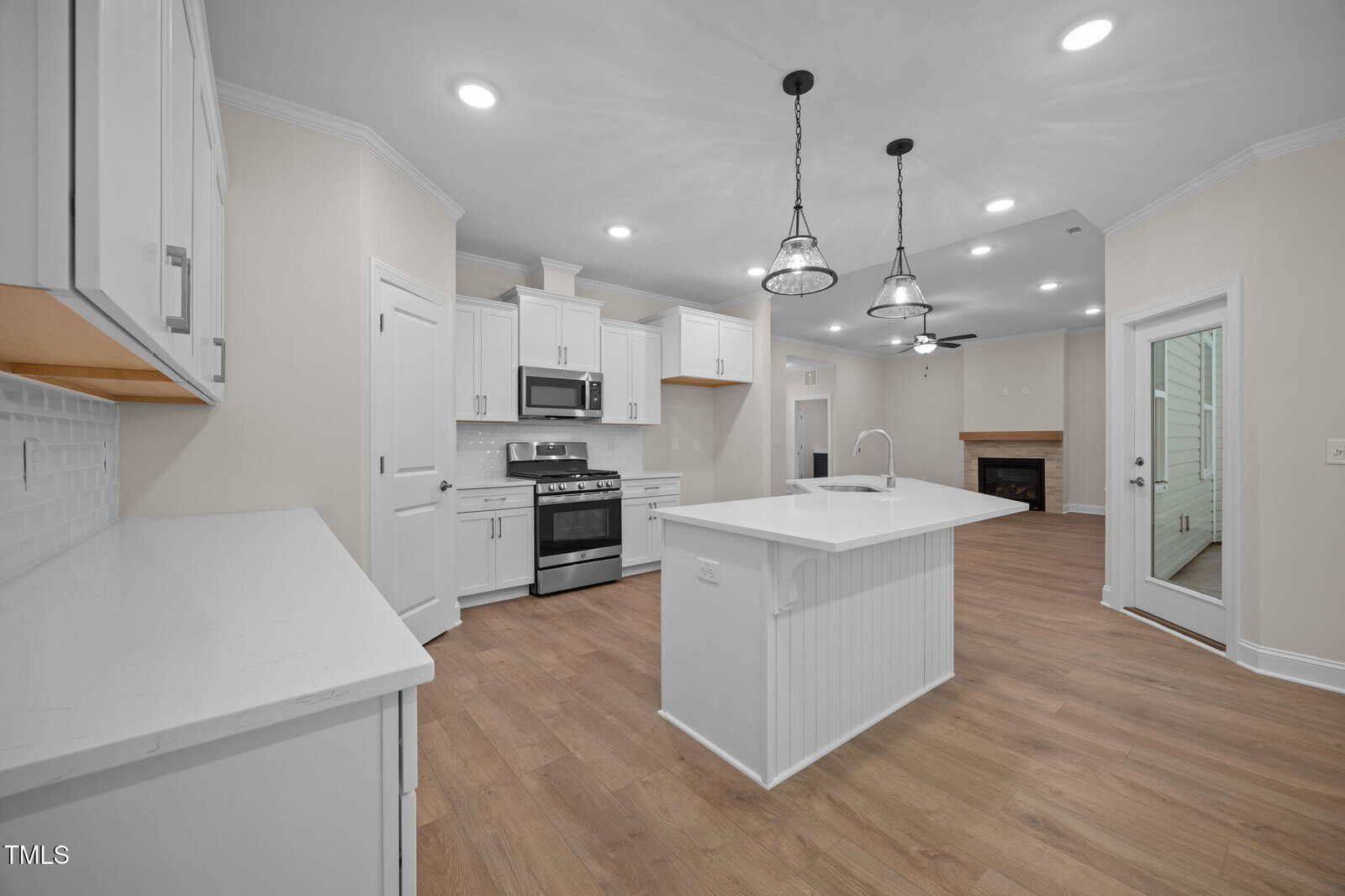 A spacious, modern kitchen with white cabinets, stainless steel appliances, a central island with a sink, wood floors, and pendant lights. The kitchen opens to a living area with a fireplace and a glass door to the outside.