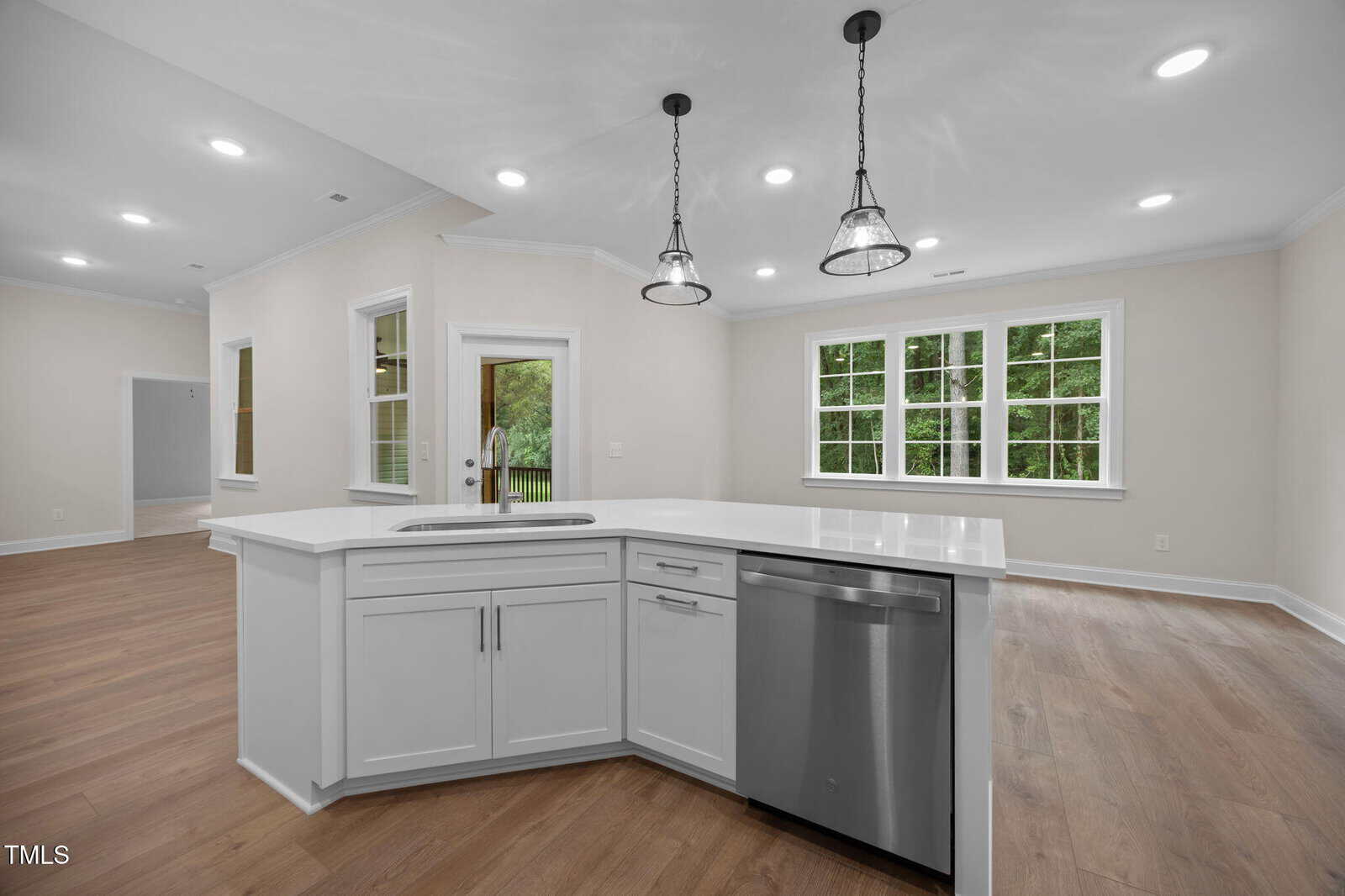 Modern kitchen with white cabinets and countertops, stainless steel appliances, pendant lights above the island, and an open layout connecting to a bright living area with large windows and wood flooring.