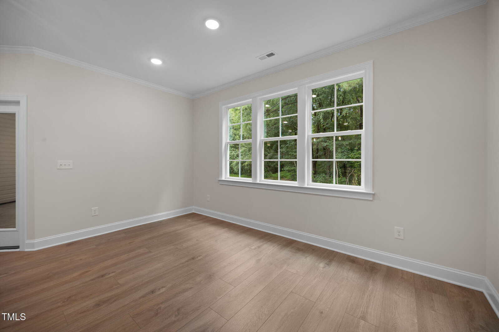 A bright, empty room with light beige walls, wood flooring, white trim, recessed ceiling lights, and a large window overlooking green trees outside.