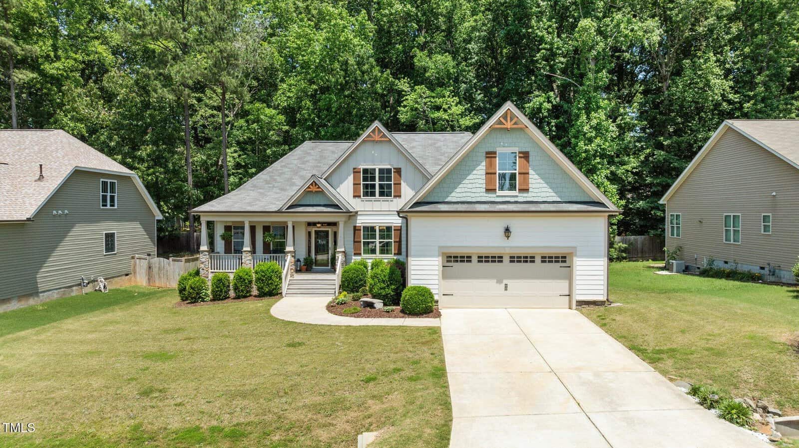 A two-story suburban house with a front porch, attached garage, manicured lawn, shrubs, and a curved walkway, surrounded by trees and neighboring homes.