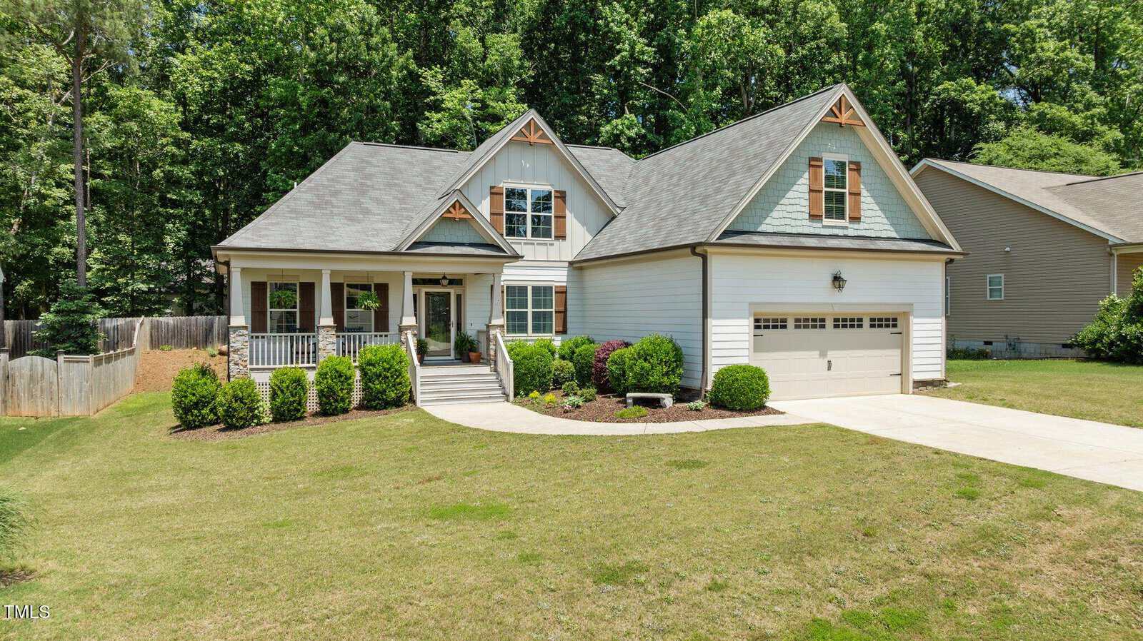 A light-gray house with a front porch, gable roof, two-car garage, and manicured lawn with shrubs and bushes, surrounded by trees and neighboring homes.