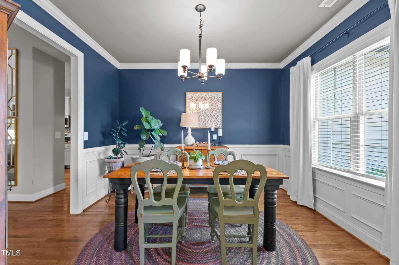 A dining room with blue walls, white wainscoting, a wooden table with four green chairs, a round rug, potted plants, a lamp on the table, a framed artwork, a chandelier, and a large window with white curtains.