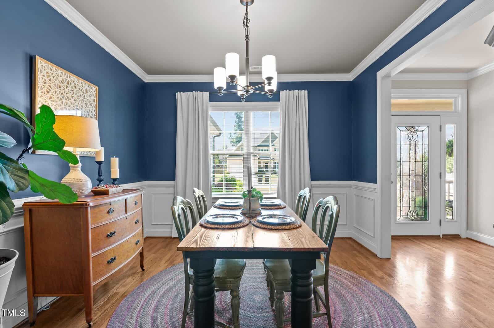 A dining room with blue walls, white trim, and a wooden table set for four. A sideboard with a lamp and decor sits on the left, and large windows with white curtains let in natural light.