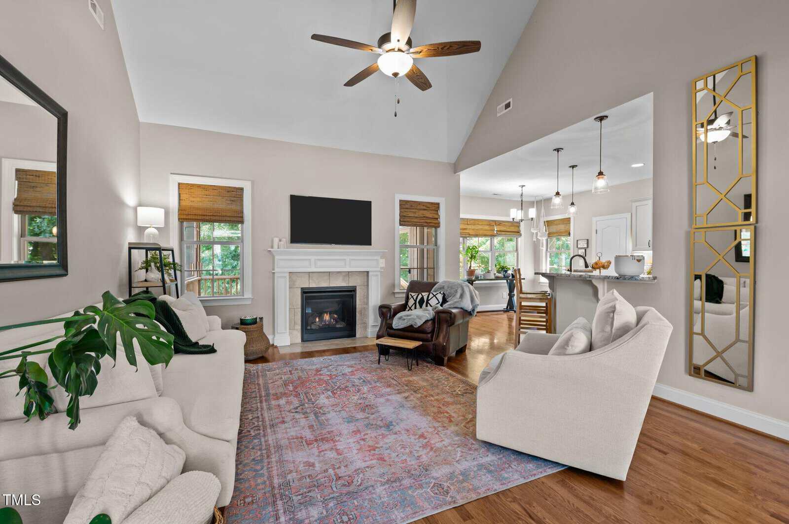 A living room with a fireplace, TV, and ceiling fan. There are two white sofas, an armchair, a colorful rug, and a large mirror. The space opens to a kitchen and dining area with hardwood floors and neutral decor.