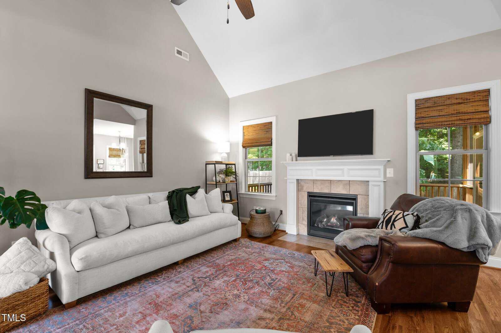 A living room with a white sofa, brown leather armchair, patterned rug, fireplace with a TV above it, large mirror, window with blinds, and a potted plant; neutral wall color and wooden accents.