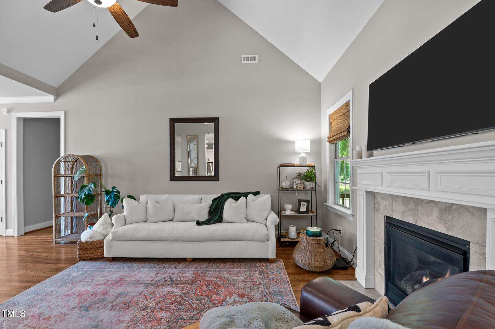 A living room with a white sofa, mirror above, rug, fireplace with TV, houseplants, a shelf with decor, and a window with a woven shade. The walls are painted light gray and the ceiling is vaulted with a ceiling fan.
