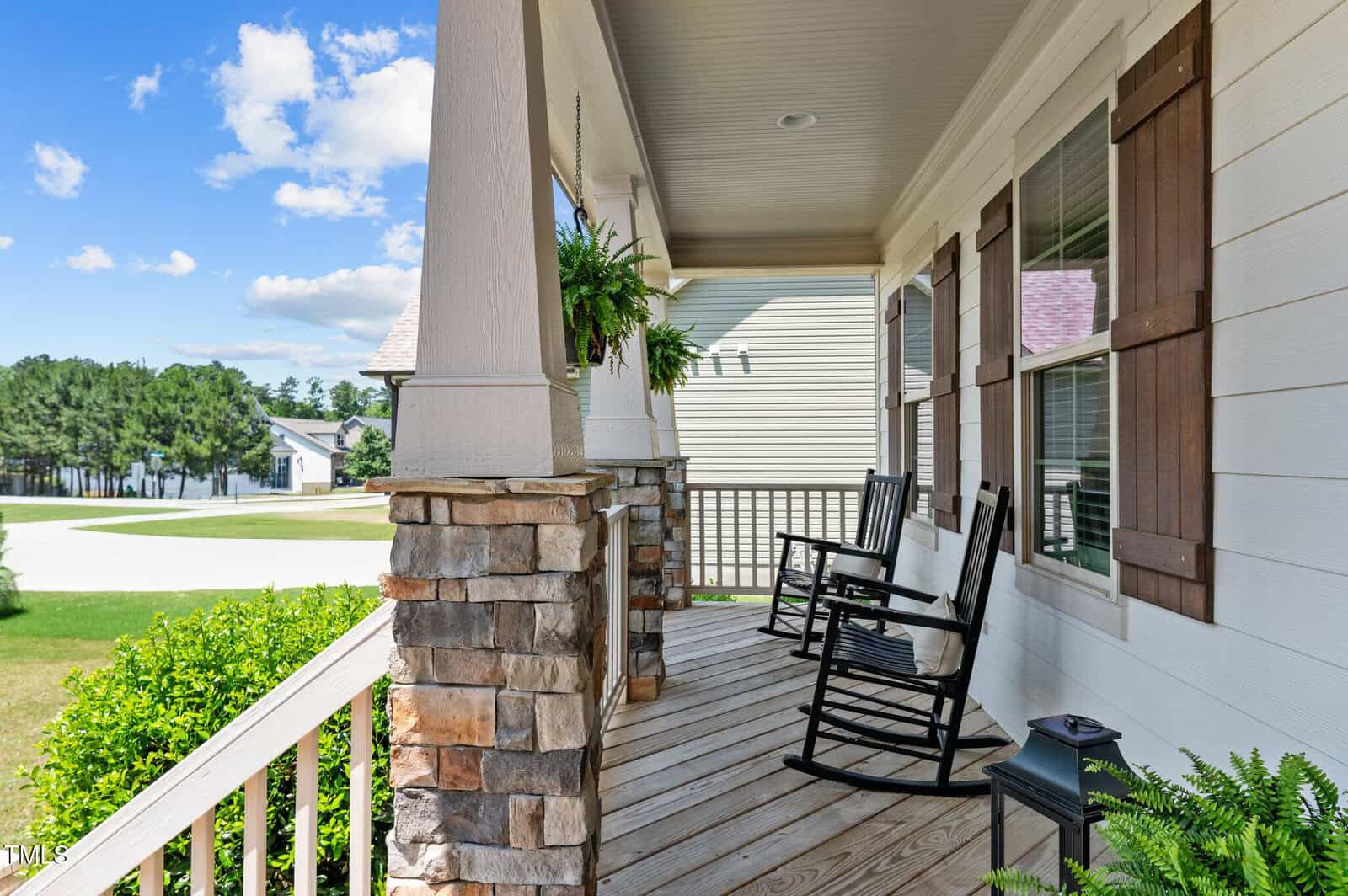 A cozy front porch with two black rocking chairs, stone pillars, wooden railings, and a hanging fern; the porch overlooks a green lawn, trees, and nearby houses under a blue sky with clouds.