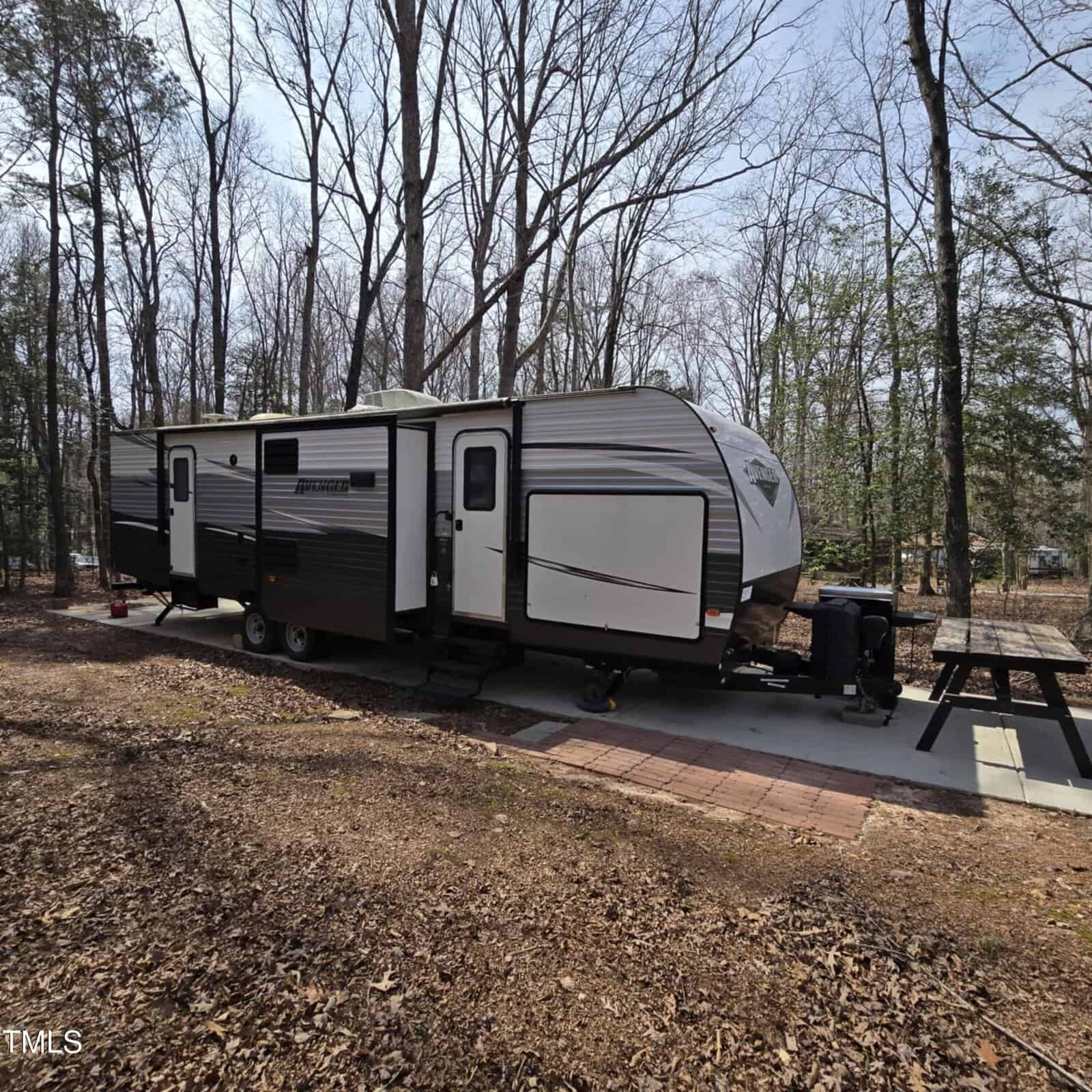 A large gray and black travel trailer is parked on a concrete pad in a wooded area, with bare trees surrounding it. A picnic table sits nearby on the pad, and fallen leaves cover the ground.