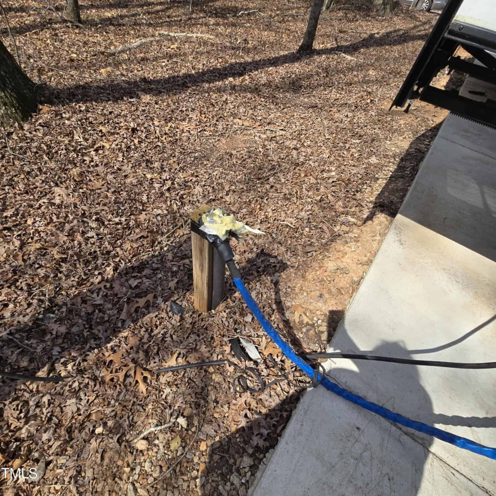 A blue hose is connected to a wooden post outdoors, with yellow insulation material around the connection. The post is surrounded by dry leaves and trees, next to a concrete surface in a wooded area.