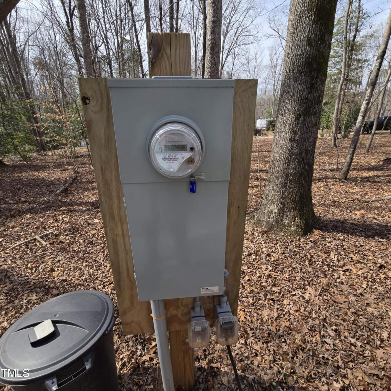 A gray outdoor electric meter box mounted on a wooden post in a wooded area with fallen leaves. Two electrical outlets are below the box, and a black trash can is nearby. Trees and a vehicle are visible in the background.