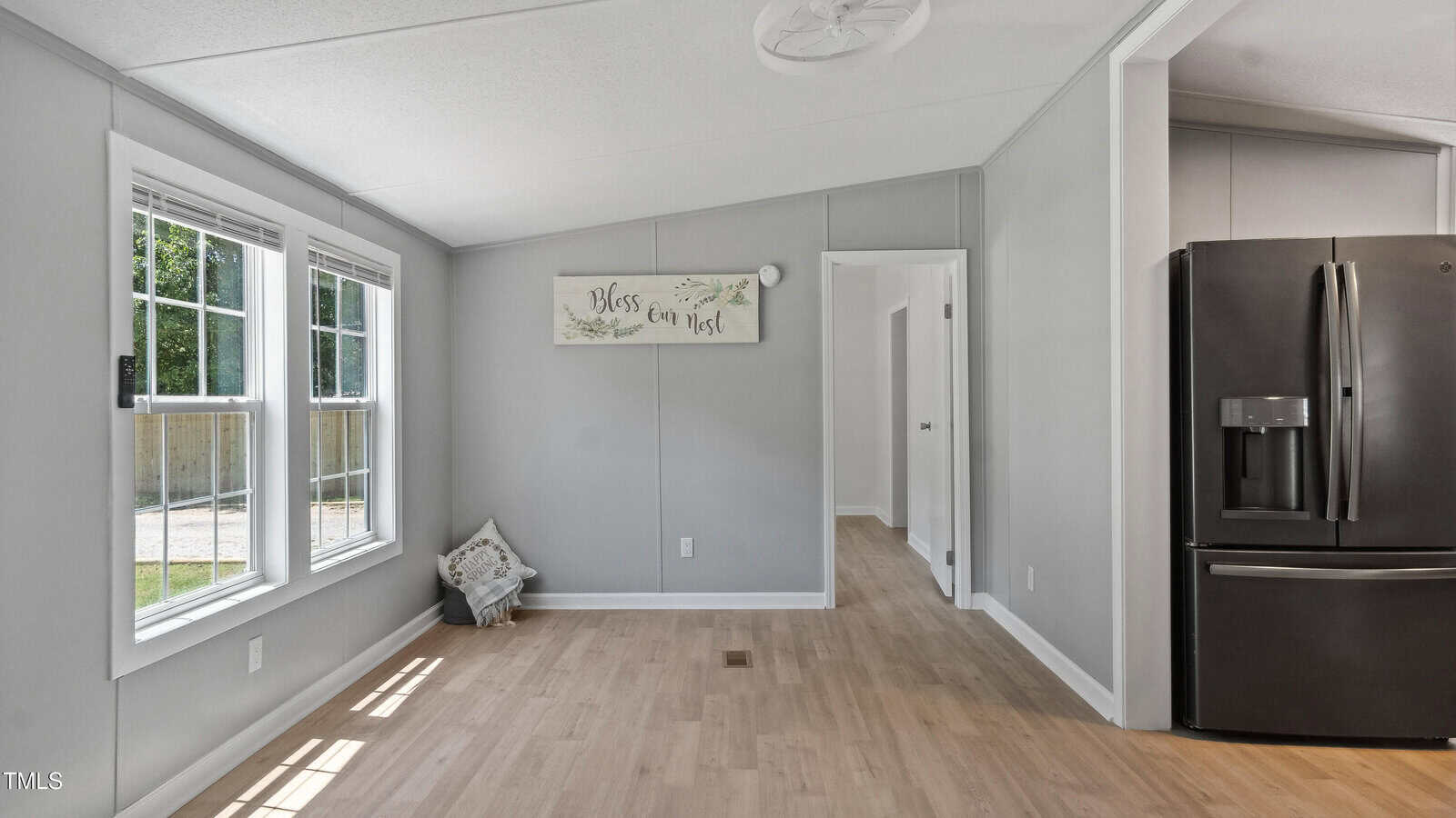 A bright room with light wood floors, gray walls, and two large windows. There is a pillow on the floor, a sign reading "Bless our nest" on the wall, and a black refrigerator in the corner near an open doorway.